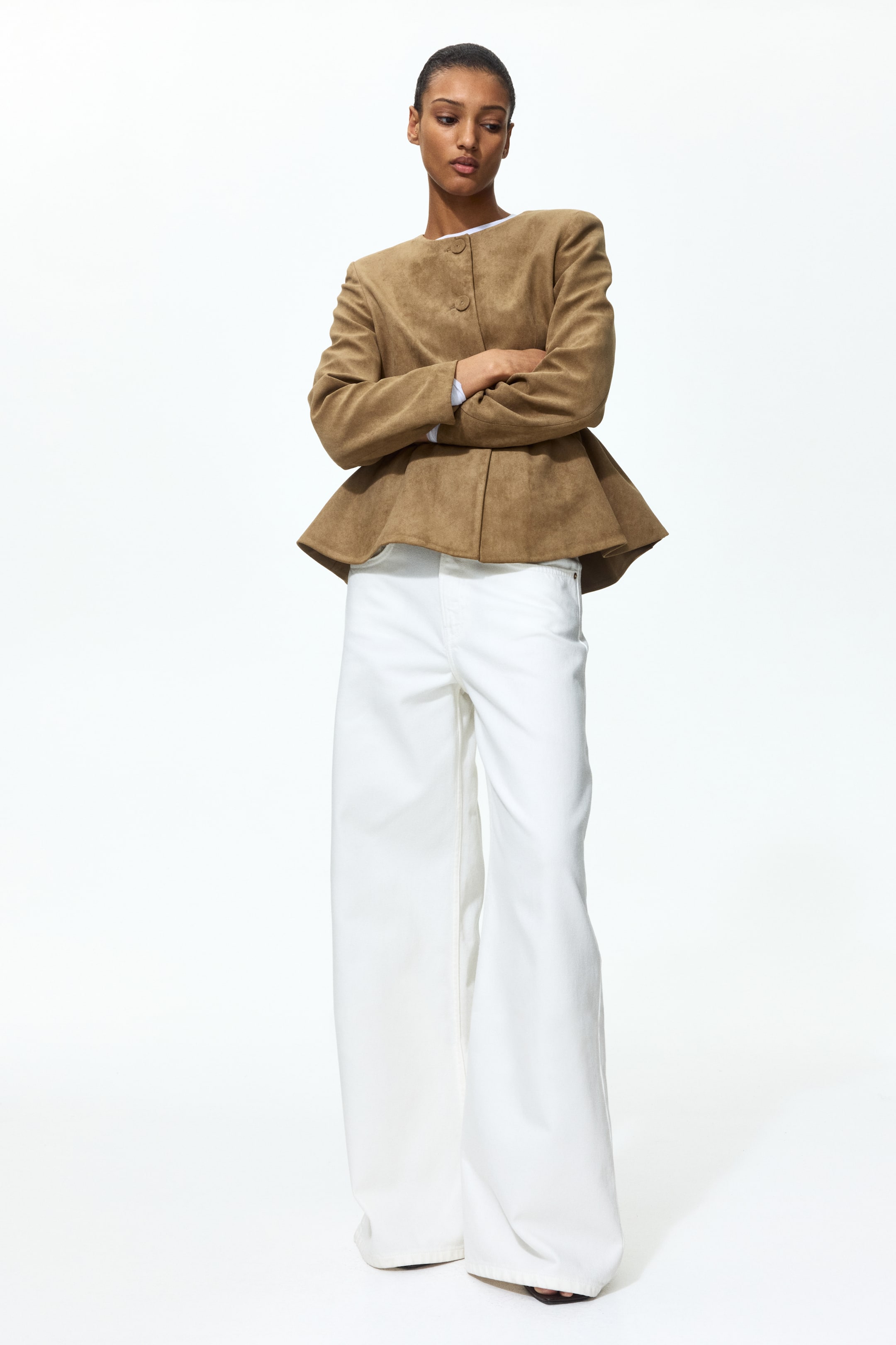 View larger image: A brown woven jacket with a round neckline and covered buttons down the front is worn by the model. It features shaping darts, a gathered seam at the waist, and a flared peplum. The model is crossing her arms, wearing it over a white top and white wide-leg trousers.