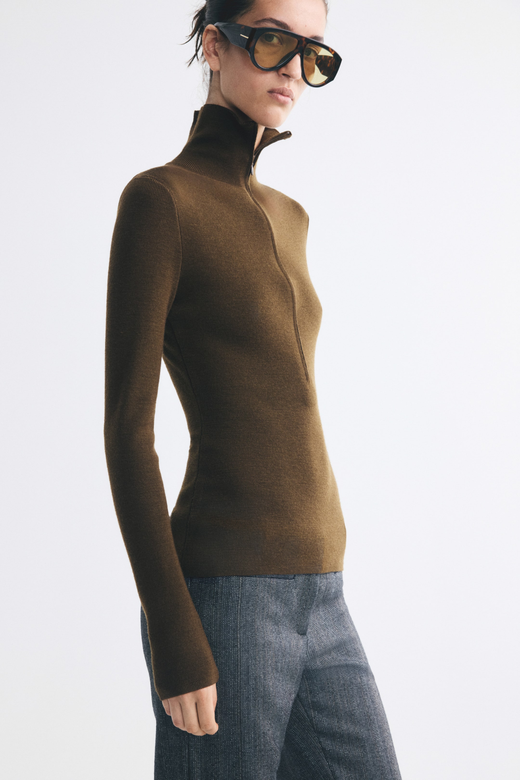View larger image: A fitted brown rib-knit top featuring a high neck with a partially unzipped front and long sleeves is worn by the model. The top is styled with grey textured trousers.