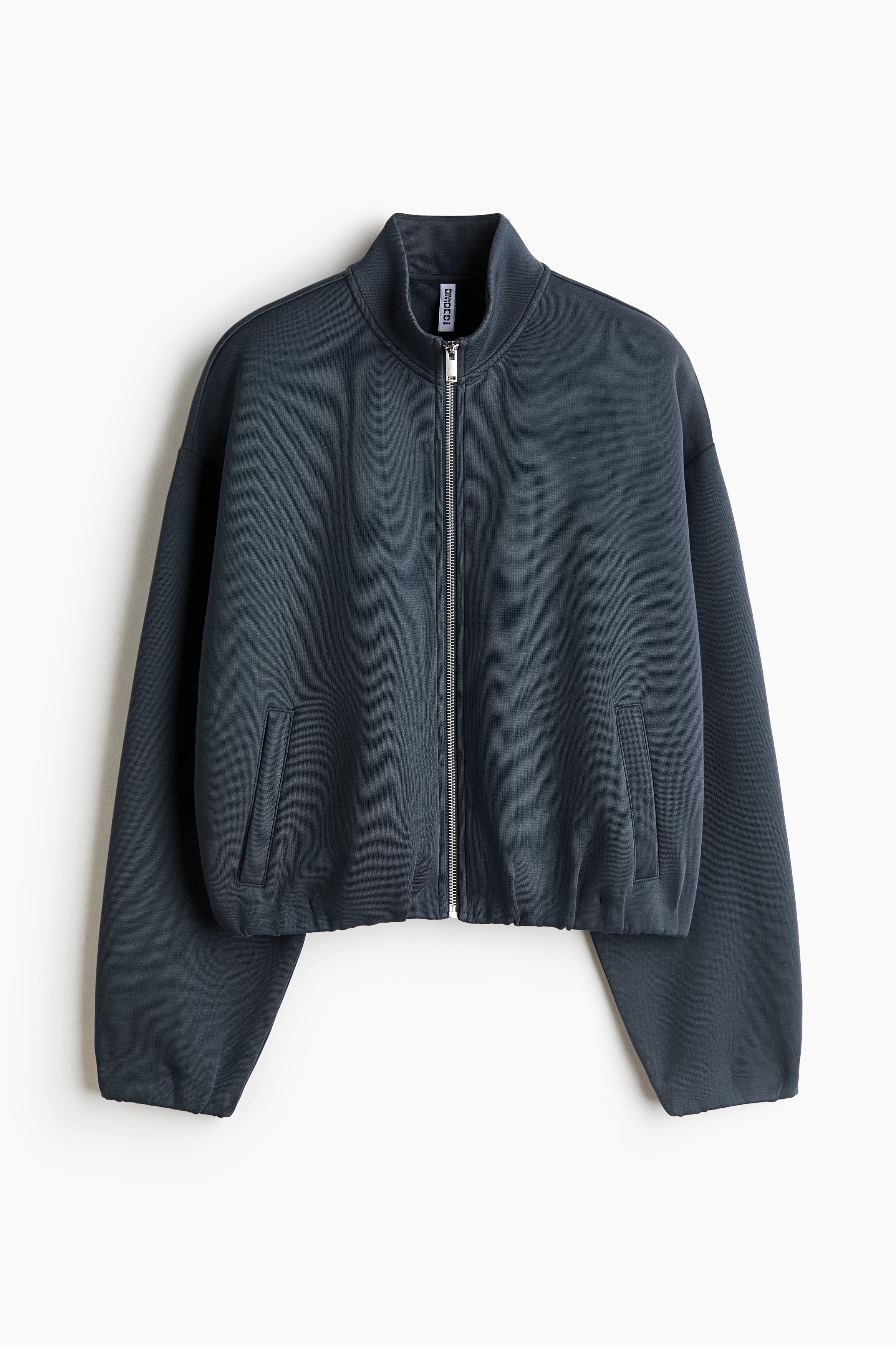 A loose-fit, dark grey scuba fabric top is shown from the front, featuring a stand collar, a metallic front zip, dropped shoulders, and long sleeves with elasticated cuffs. The top is styled with matching wide-leg trousers, highlighting its relaxed silhouette and elasticated hem.