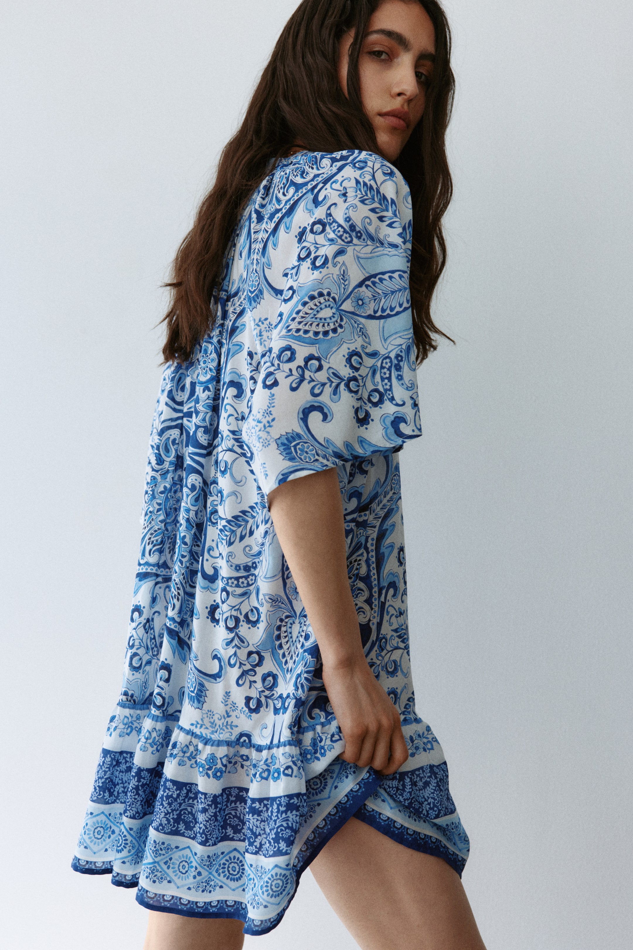 Größeres Bild ansehen: Das Kleid präsentiert eine lockere, fließende Silhouette mit kurzen, weiten Ärmeln, die von der Seite nach hinten abgewinkelt sind. Es zeigt ein aufwendiges blau-weißes Paisley-Muster, und der gestufte Rüschen-Saum endet oberhalb des Knies und offenbart ein inneres Muster, wenn er sanft angehoben wird.