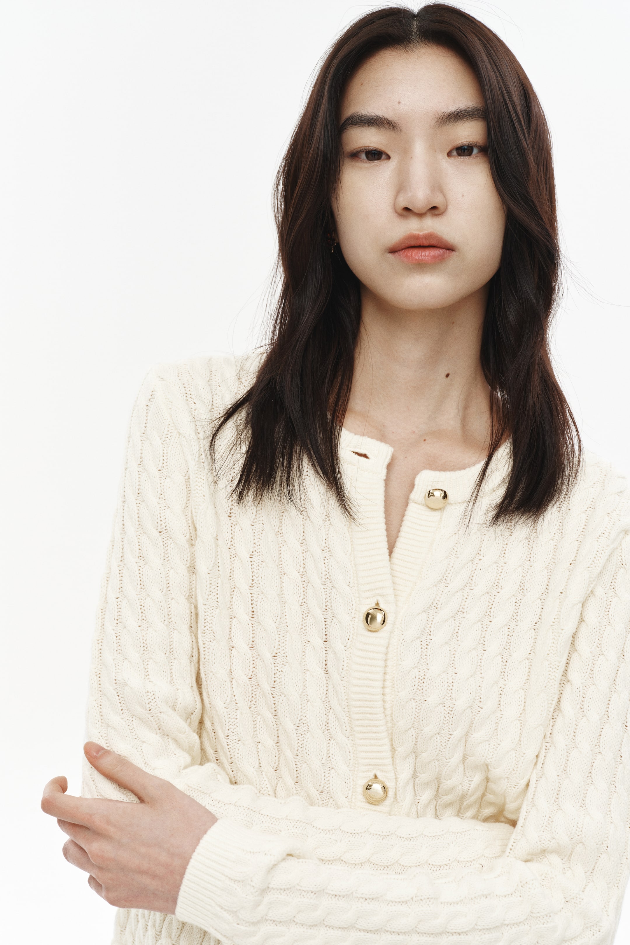 이미지 확대 보기: 크림색 케이블 니트 가디건이 약간 왼쪽을 향해 입혀져 있으며, V넥 라인과 앞면을 여미는 골드 톤의 둥근 버튼이 특징입니다. 몸통과 긴 소매 전체에 걸쳐 텍스처가 있는 케이블 니트 패턴이 눈에 띄며, 소매 끝은 리브드 커프로 마무리되어 있습니다.