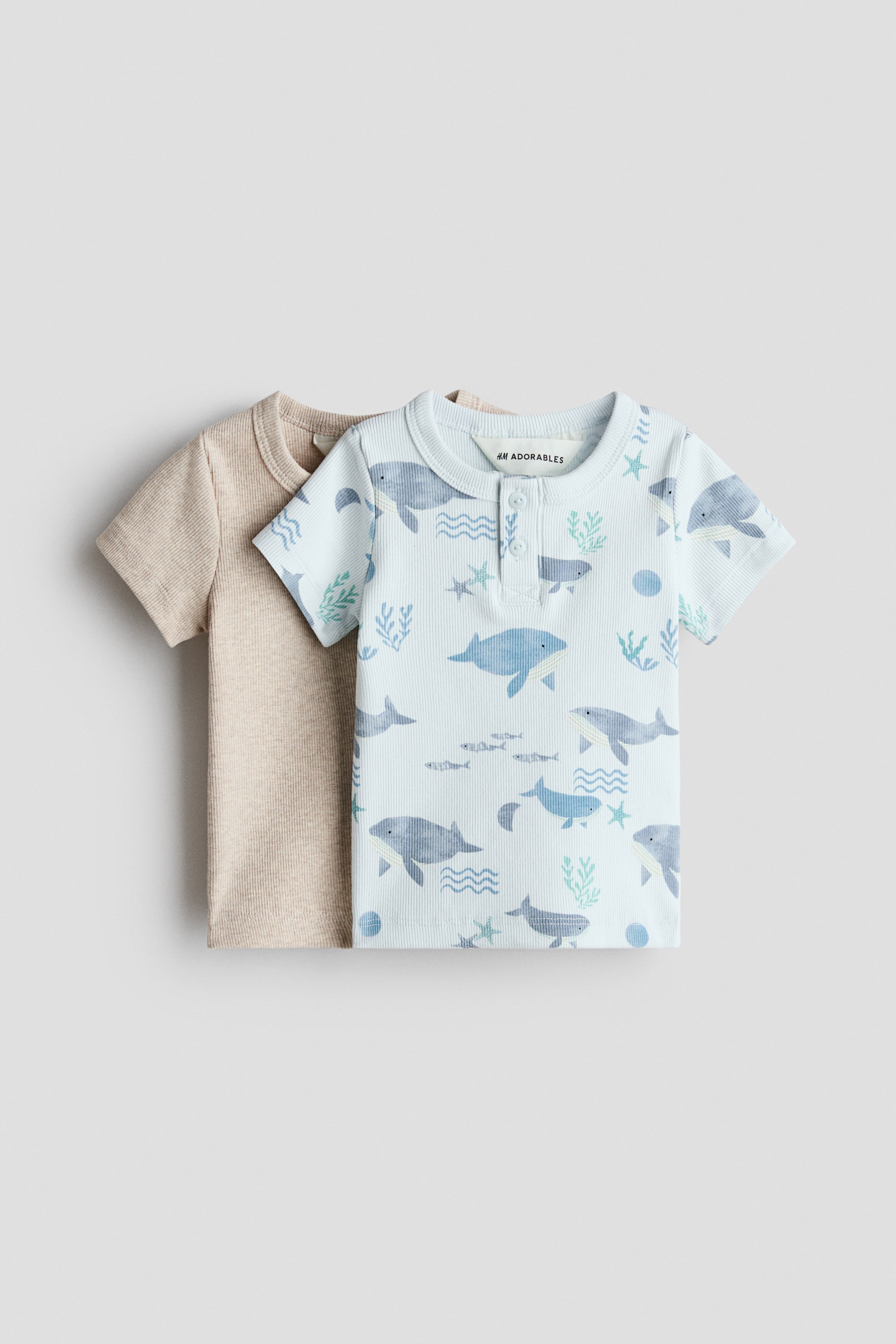 Größeres Bild ansehen: Zwei kurzärmelige Tops von H&M Adorables sind von vorne zu sehen. Das vordere Top ist hellblau und zeigt eine gerippte Struktur sowie ein ozeanisches Muster aus Walen und Fischen. Es hat einen runden Ausschnitt und eine Knopfleiste mit zwei Knöpfen. Ein einfarbiges, hellbeiges, geripptes Kurzarm-Top ist teilweise dahinter sichtbar.