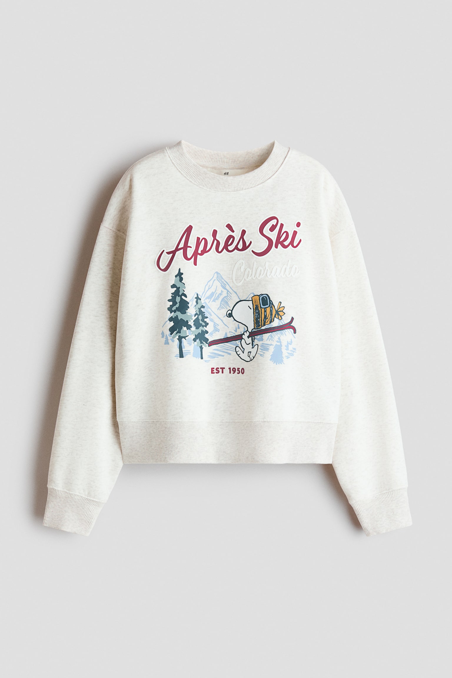 An off-white crewneck sweatshirt, shown from the front, features drop shoulders, ribbed cuffs, and a wide ribbed hem. The chest displays a graphic with red cursive text reading "Aprï¿½s Ski," the word "Colorado" in light blue, and an illustration of the cartoon character Snoopy carrying skis in a snowy mountain landscape.