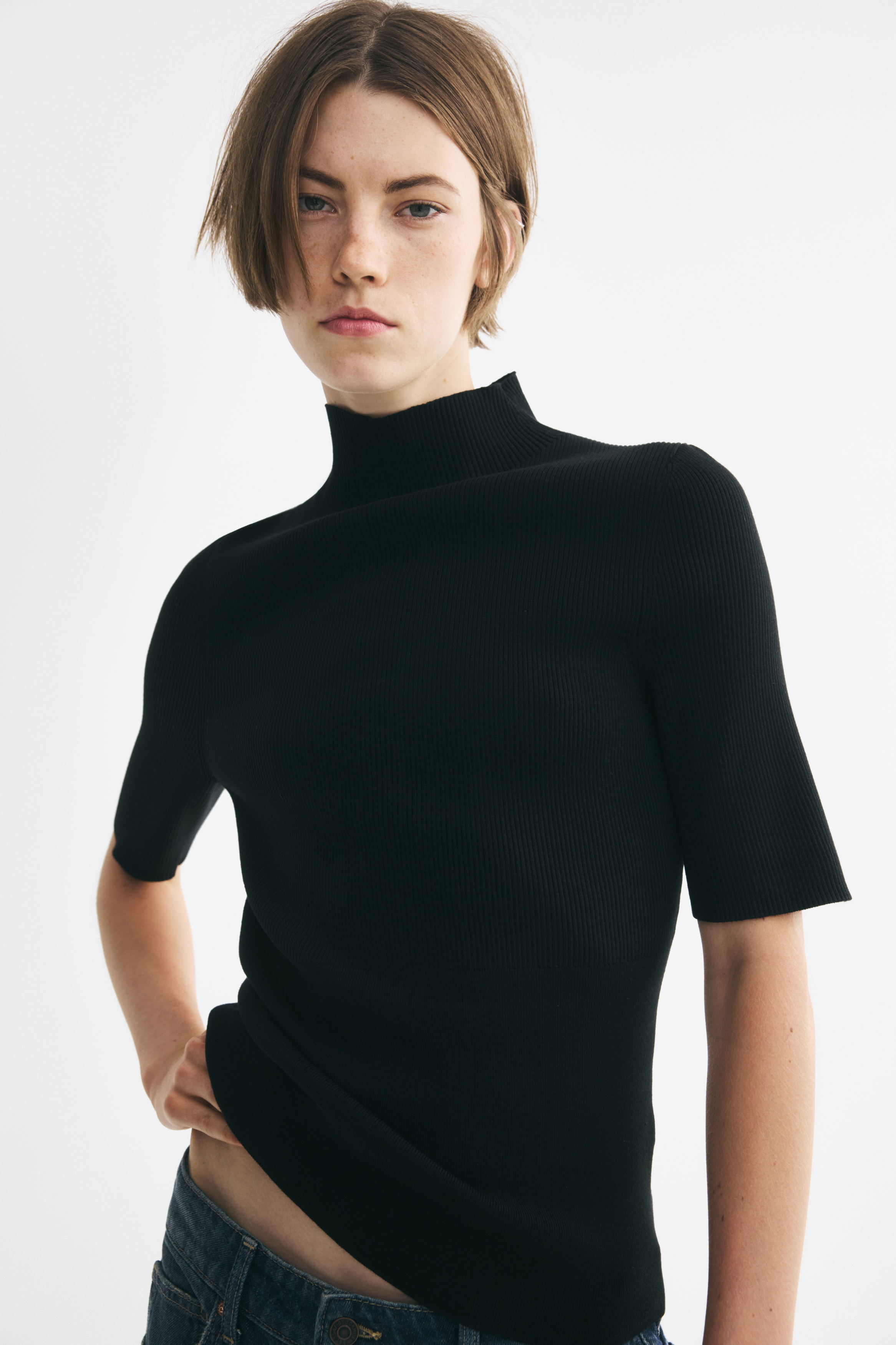 Größeres Bild ansehen: Ein schwarzes, figurbetontes Top aus dichtem Rippstrick wird von vorne gezeigt und verfügt über einen hohen Stehkragen sowie kurze Ärmel. Das eng anliegende Top liegt über dem Bund einer blauen Jeans, wobei der gerade Saum auf einer Seite leicht angehoben ist.