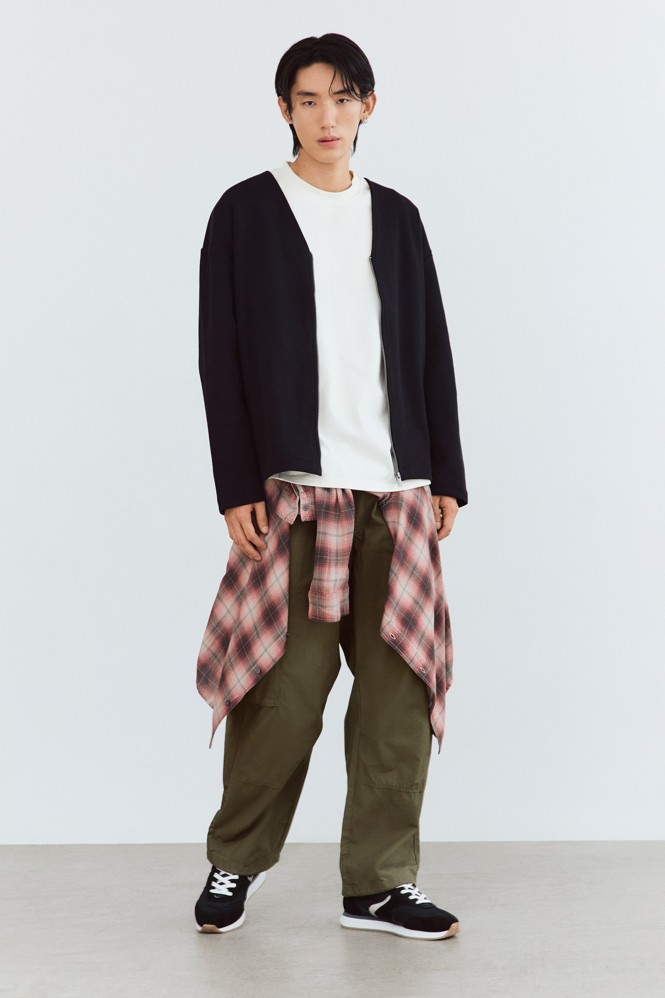 View larger image: The model is wearing olive green cargo pants in a relaxed fit, featuring visible knee pleats and flap side pockets. The pants are styled with a plaid shirt tied around the waist, a white T-shirt, a black cardigan, and black sneakers.