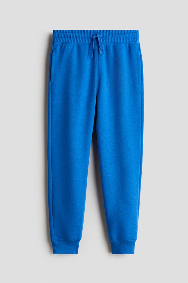 Se muestran pantalones de chándal azules desde el FRENTE, con una cintura elástica con cordón, bolsillos laterales visibles y piernas ligeramente ajustadas con dobladillos acanalados. - 1