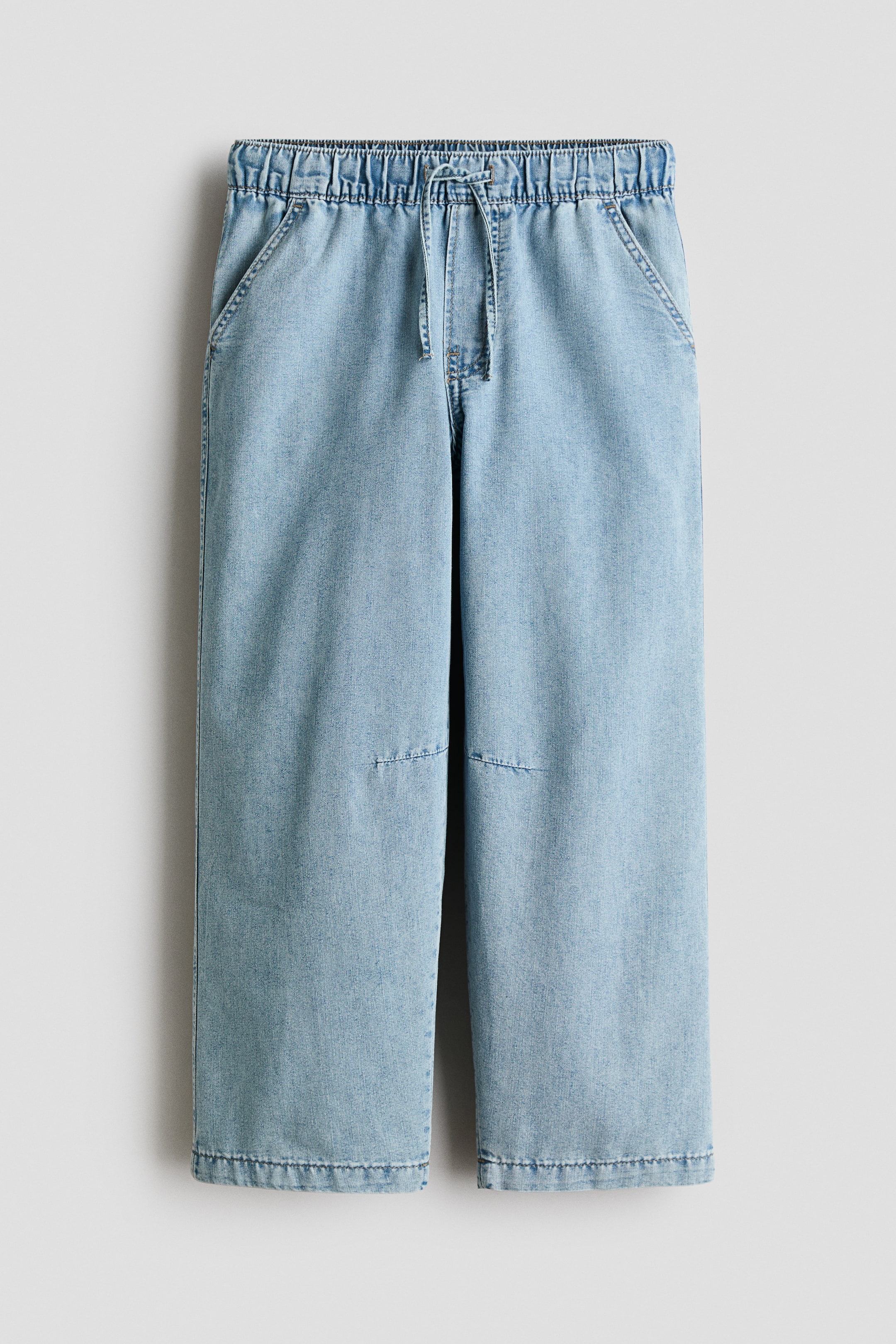 Größeres Bild ansehen: Hellblaue Denim-Hosen werden von vorne gezeigt, mit einem elastischen Kordelzugbund, schrägen Vordertaschen und weiten Beinen mit rechteckigen Patch-Details an den Knien und genähten Säumen.
