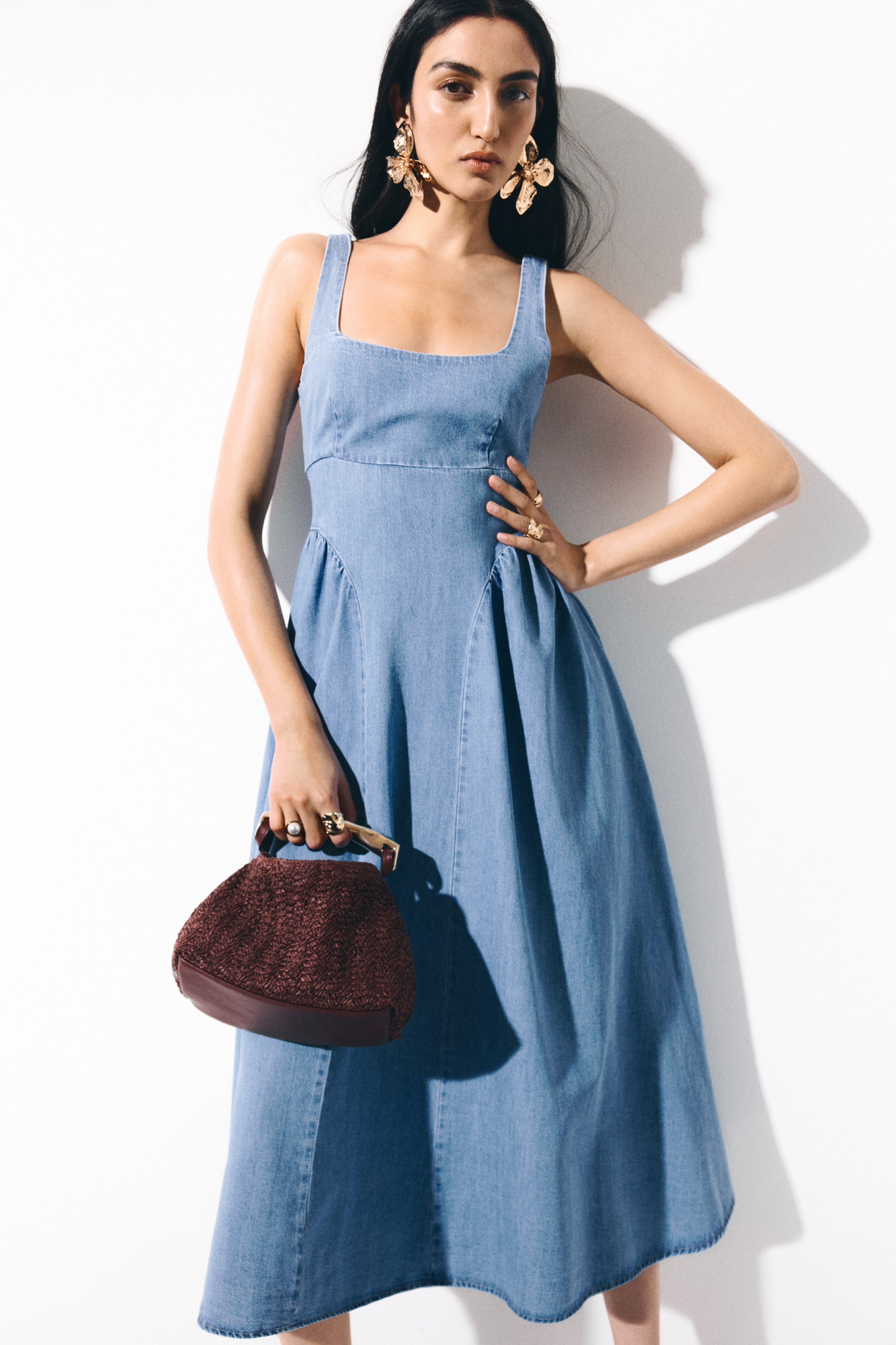 View larger image: A light blue denim dress showcasing a square neckline, wide straps, and a fitted bodice. It transitions into an A-line midi skirt with vertical seams that define its structure, slightly angled towards the front-right.