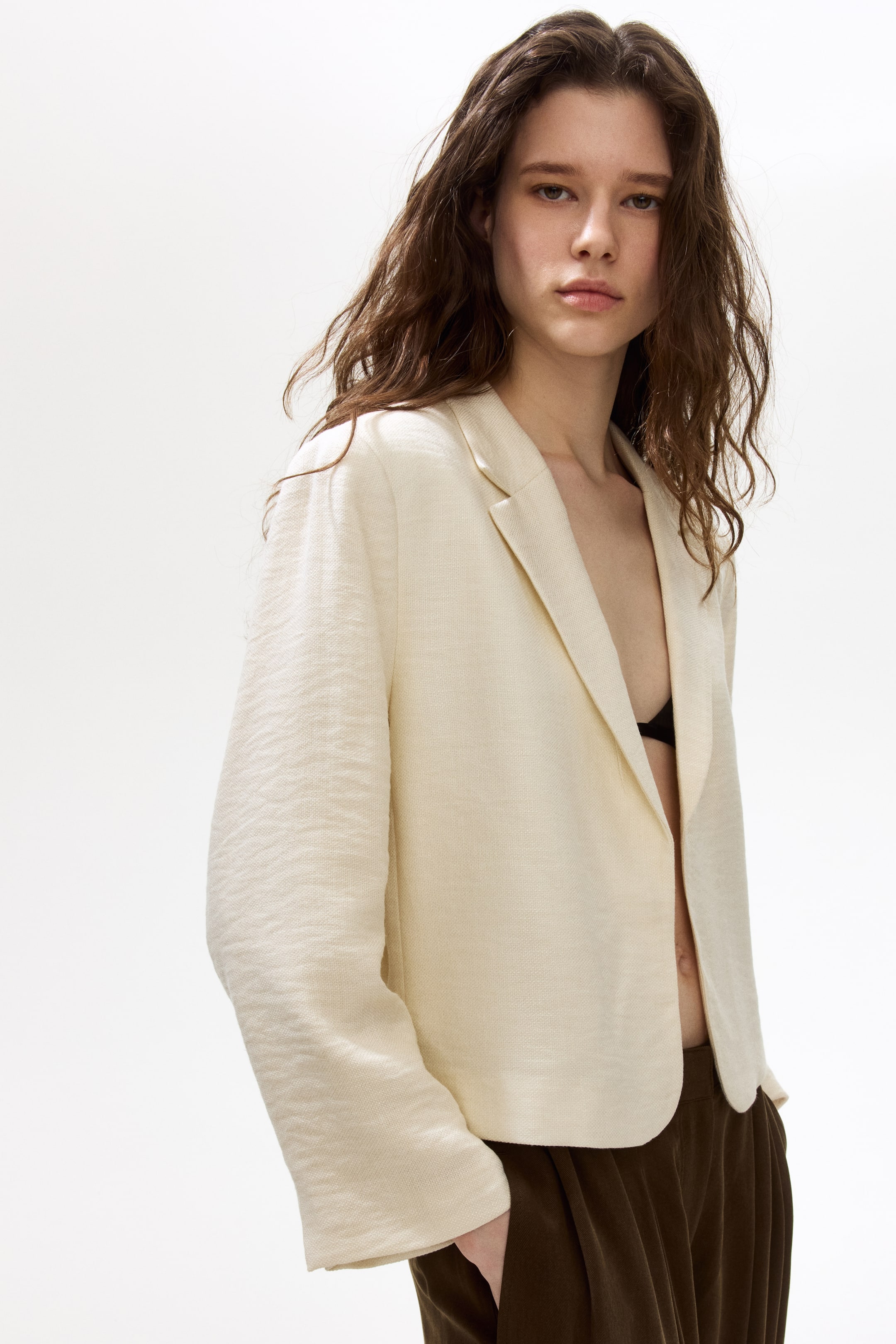 Ver imagen más grande: Una blazer de color crema presenta un frente abierto con solapas de muesca, sus mangas largas muestran una textura arrugada distintiva. La blazer se lleva sobre un sujetador negro y pantalones marrón oscuro, posicionada en un ligero ángulo frontal lateral.