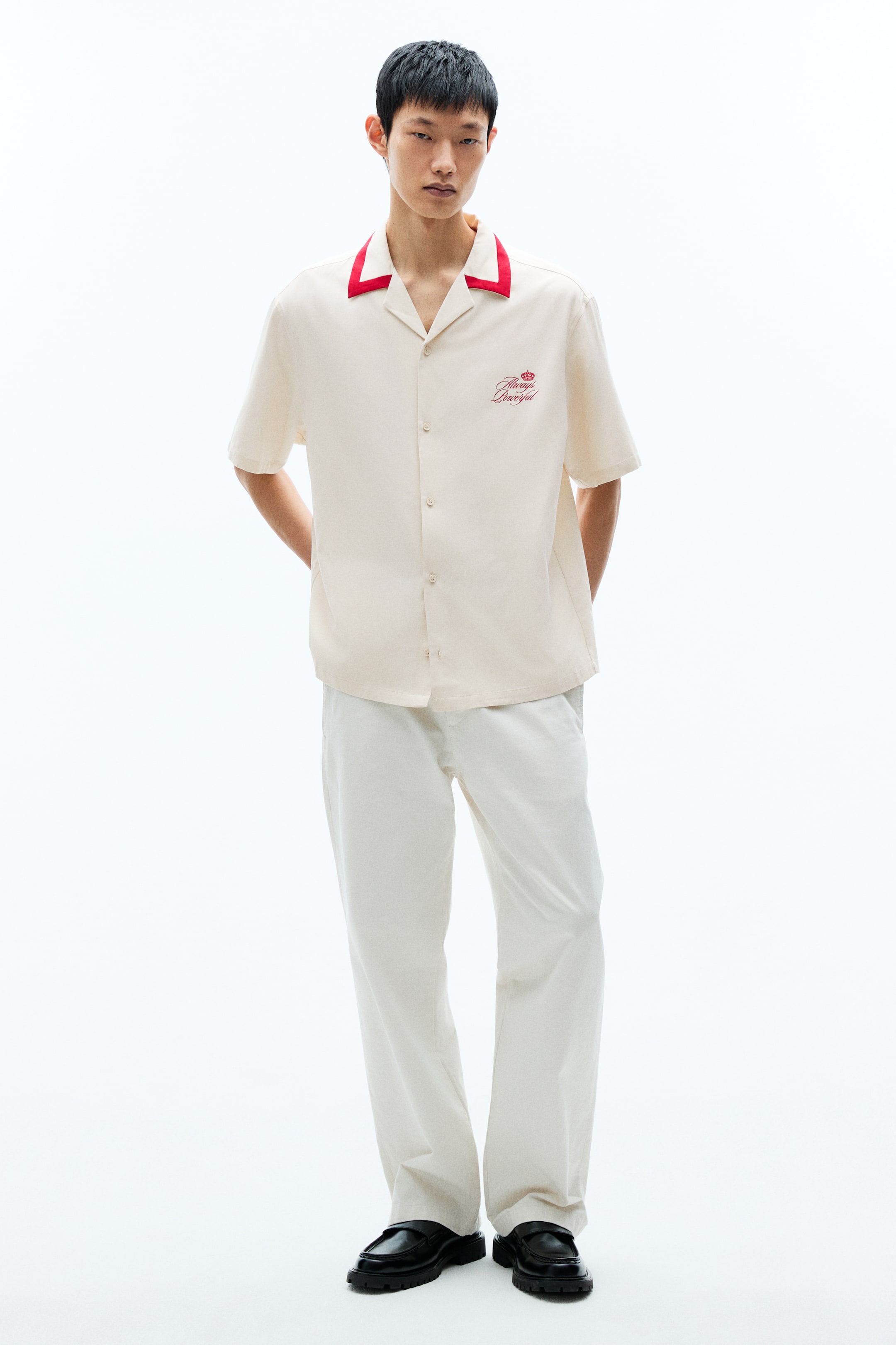 查看大圖: 奶油色短袖襯衫，飾有紅色滾邊的露營領，左胸以紅色刺繡呈現皇冠與文字，前襟中央備有一列鈕扣，正面展示。