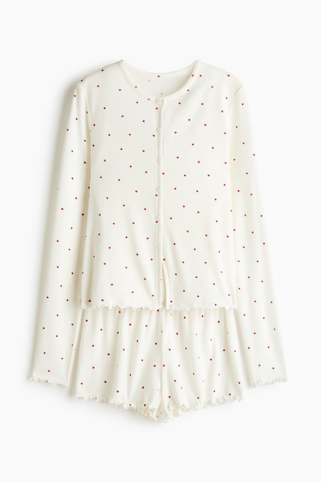 A two-piece pajama set, shown from the front, is featuring a long-sleeved button-up top and matching shorts made from an off-white fabric with an all-over print of tiny red hearts. Both pieces are displaying ruffled lettuce-edge hems along the sleeve cuffs, bottom hem, and leg openings. - 1