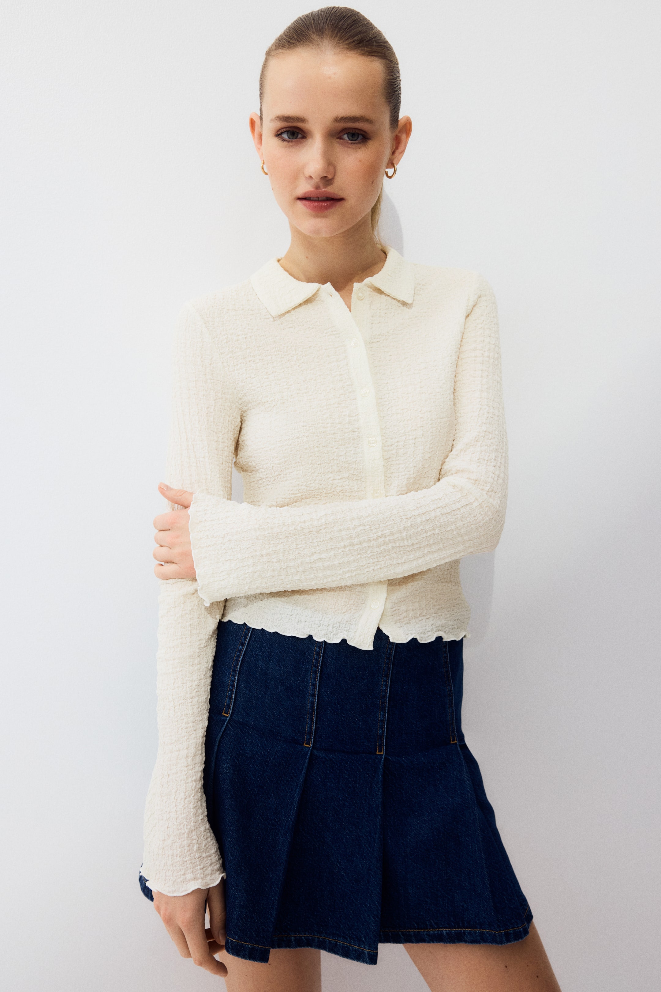 View larger image: An off-white fitted long-sleeved top showcases a distinct crinkled texture, a pointed collar, and a button-front placket with light-colored buttons. The top is displayed facing forward, highlighting a wavy hemline and subtly flared cuffs.