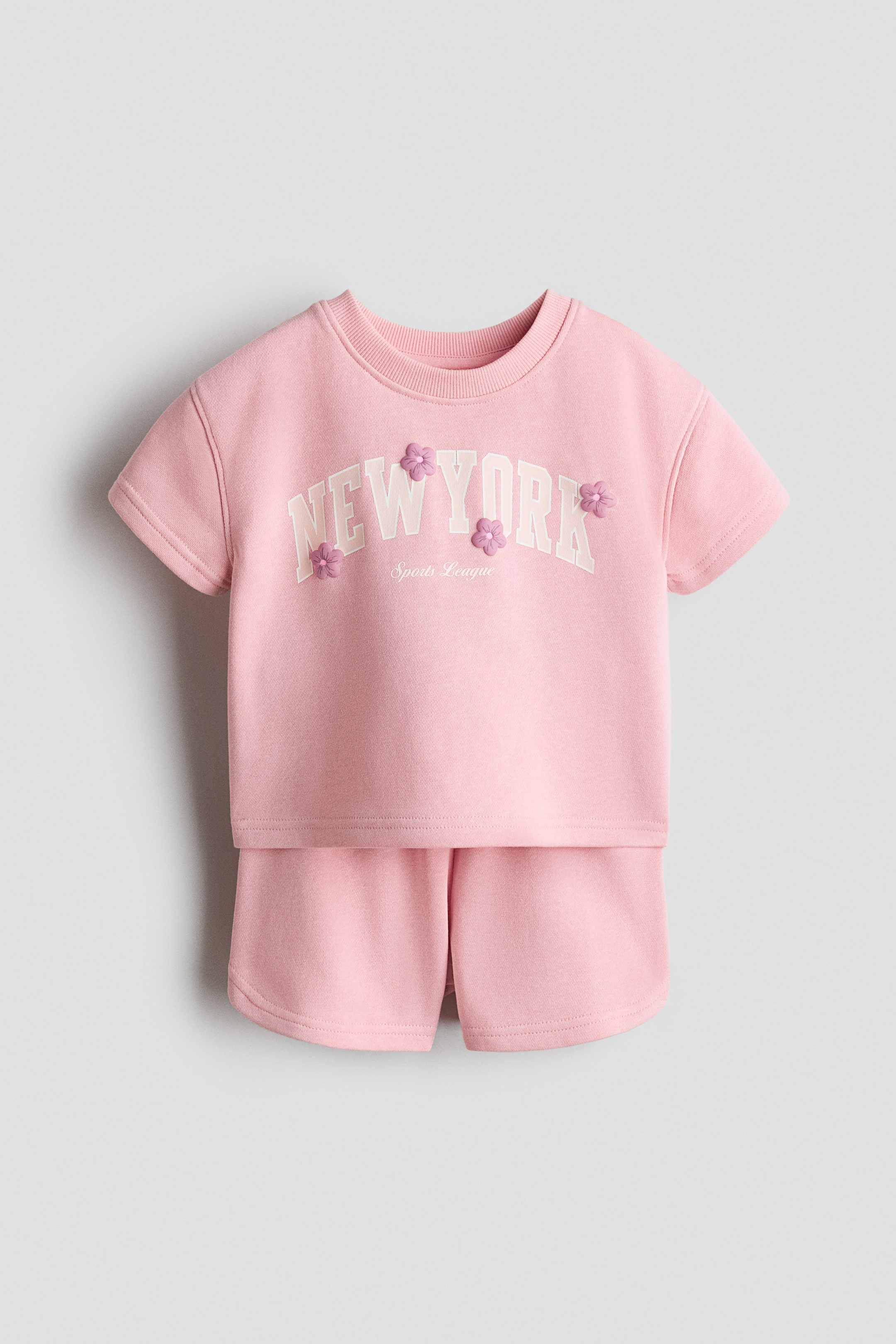 Ampliar la imagen: Se muestra de frente un conjunto compuesto por un top rosa de manga corta y shorts a juego. El top presenta un cuello redondo acanalado y el texto "NEW YORK" en blanco con un contorno tenue, realzado con pequeñas aplicaciones de flores moradas, con el texto "Sports League" debajo. Los shorts lisos color rosa están diseñados con dobladillos redondeados.