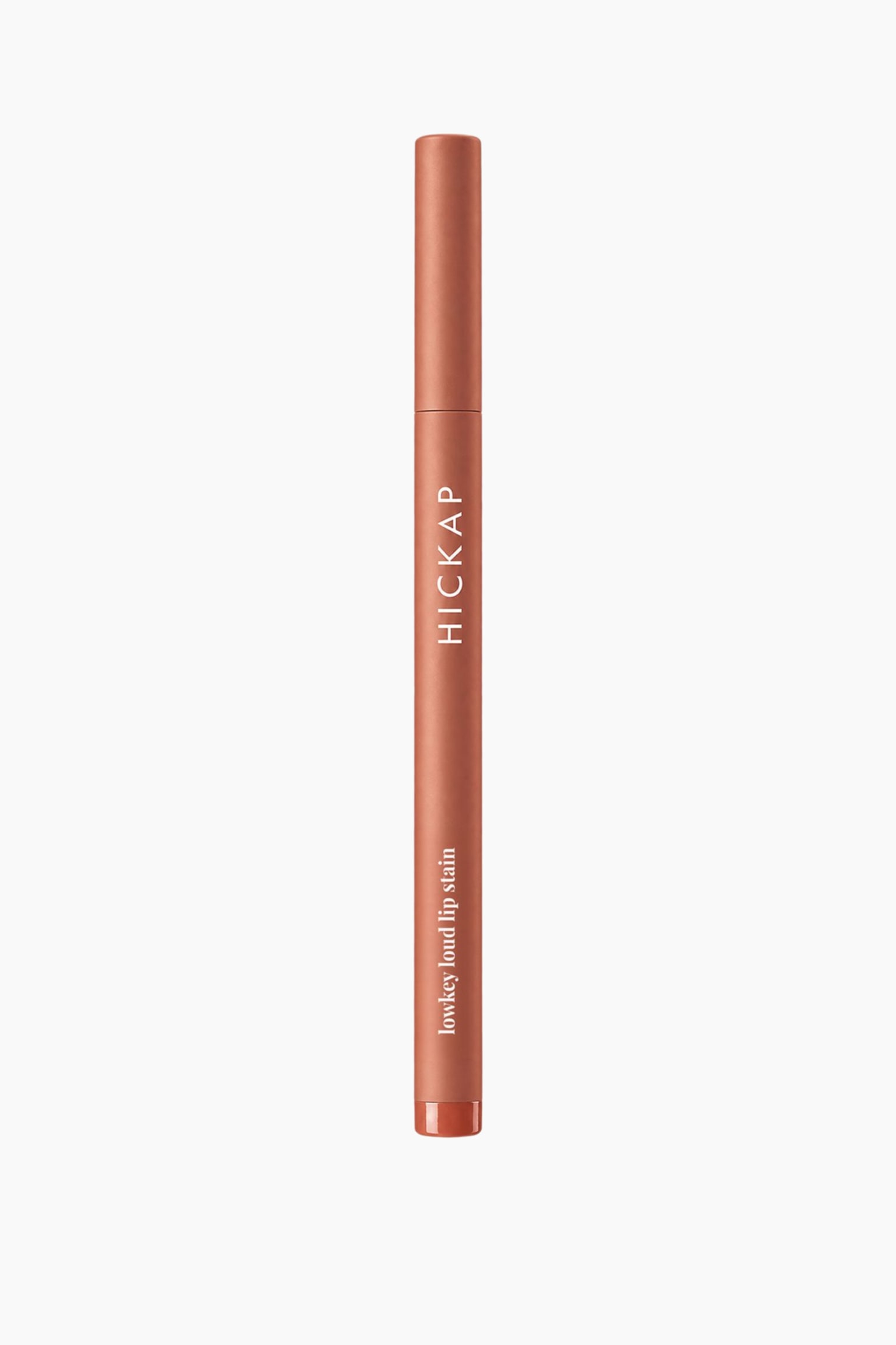 En slimmad, cylindrisk lip stain-penna står lodrätt och visar ett matt terrakottabrunt hölje med ett matchande lock. Pennan har varumärket "HICKAP" tryckt vertikalt i vita versaler på kroppen, med "lowkey loud lip stain" tryckt i vit gemener nära den glansiga basen. - 1