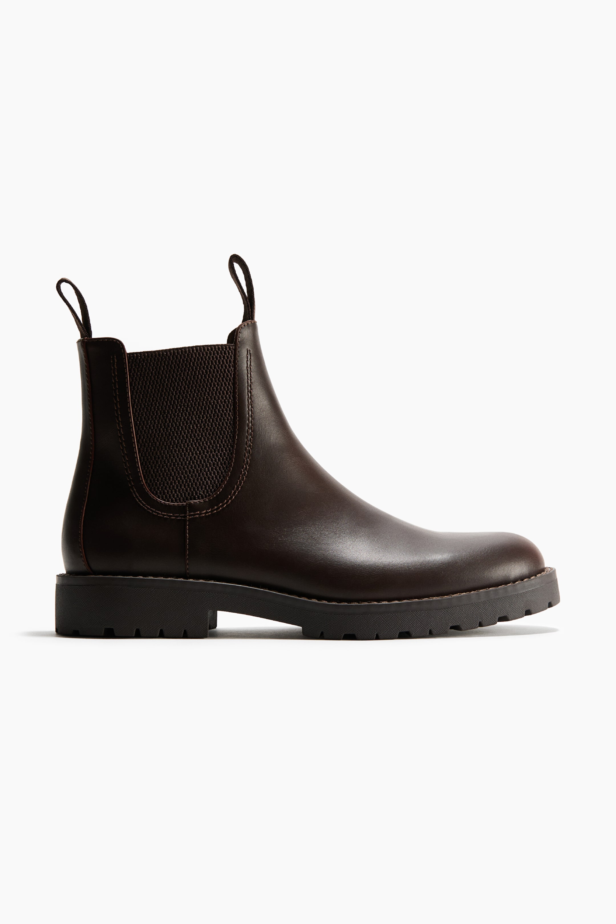 View larger image: A dark brown leather Chelsea boot is shown in side profile, featuring elastic side panels and pull tabs at the front and back of the collar. The boot sits on a thick black lug sole with a rounded toe.