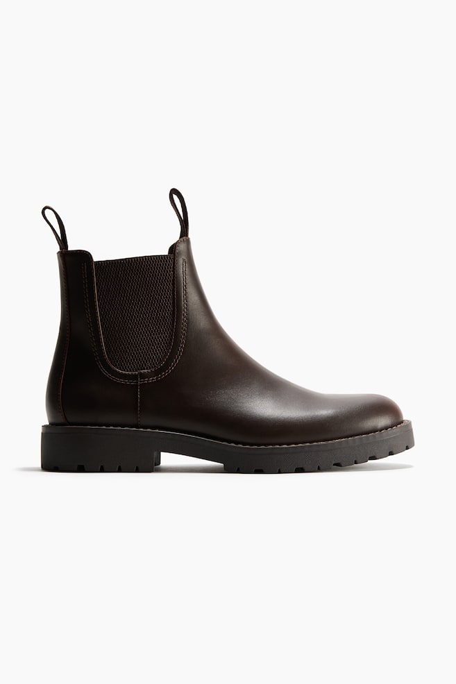 Ein dunkelbrauner Chelsea-Boot aus Leder ist in der Seitenansicht dargestellt und verfÃ¼gt Ã¼ber elastische SeiteneinsÃ¤tze sowie Zuglaschen an der Vorder- und RÃ¼ckseite des Schafts. Der Stiefel ruht auf einer robusten, schwarzen Profilsohle mit abgerundeter Zehenpartie. - 1