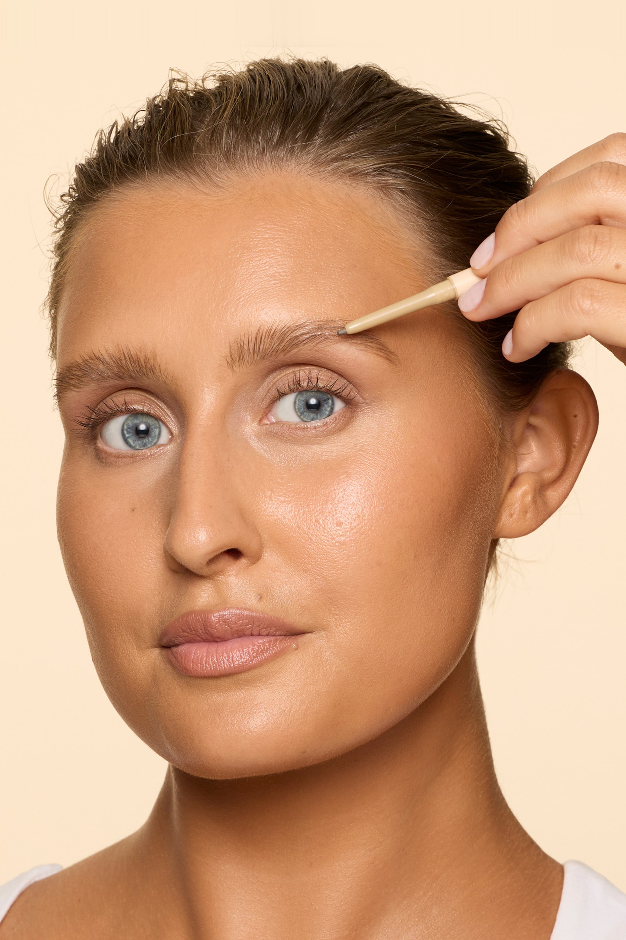 Se større billede: En slank, lysebrun øjenbrynsblyant med en gråbrun spids bliver holdt af en hånd, som påfører den på et øjenbryn. Blyanten er vinklet nedad og forbinder med øjenbrynshårene.