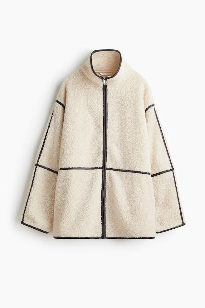 Une veste teddy de couleur crème à coupe ample est présentée de face, avec un col montant et une fermeture éclair noire au centre. Un liseré foncé en tissu enduit souligne le col, la fermeture éclair, les épaules tombantes et l'ourlet, tout en créant des lignes de design horizontales sur le corps et les manches. - 1
