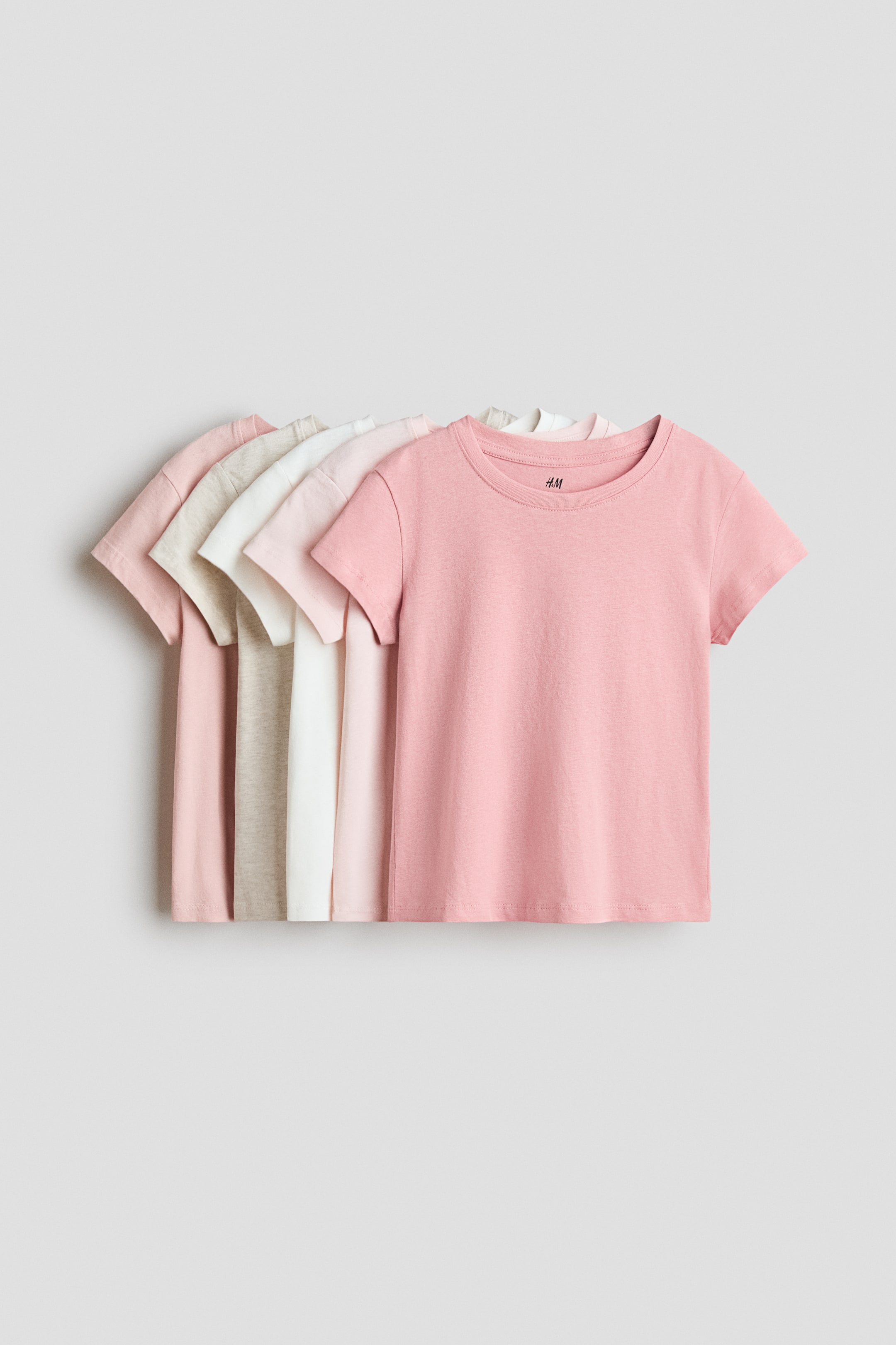 View larger image: T-shirts are displayed front-facing, featuring round, rib-trimmed necklines and short sleeves with gently dropped shoulders. They are stacked in varying light colours, including pink, white, and beige, with the frontmost pink T-shirt fully visible.