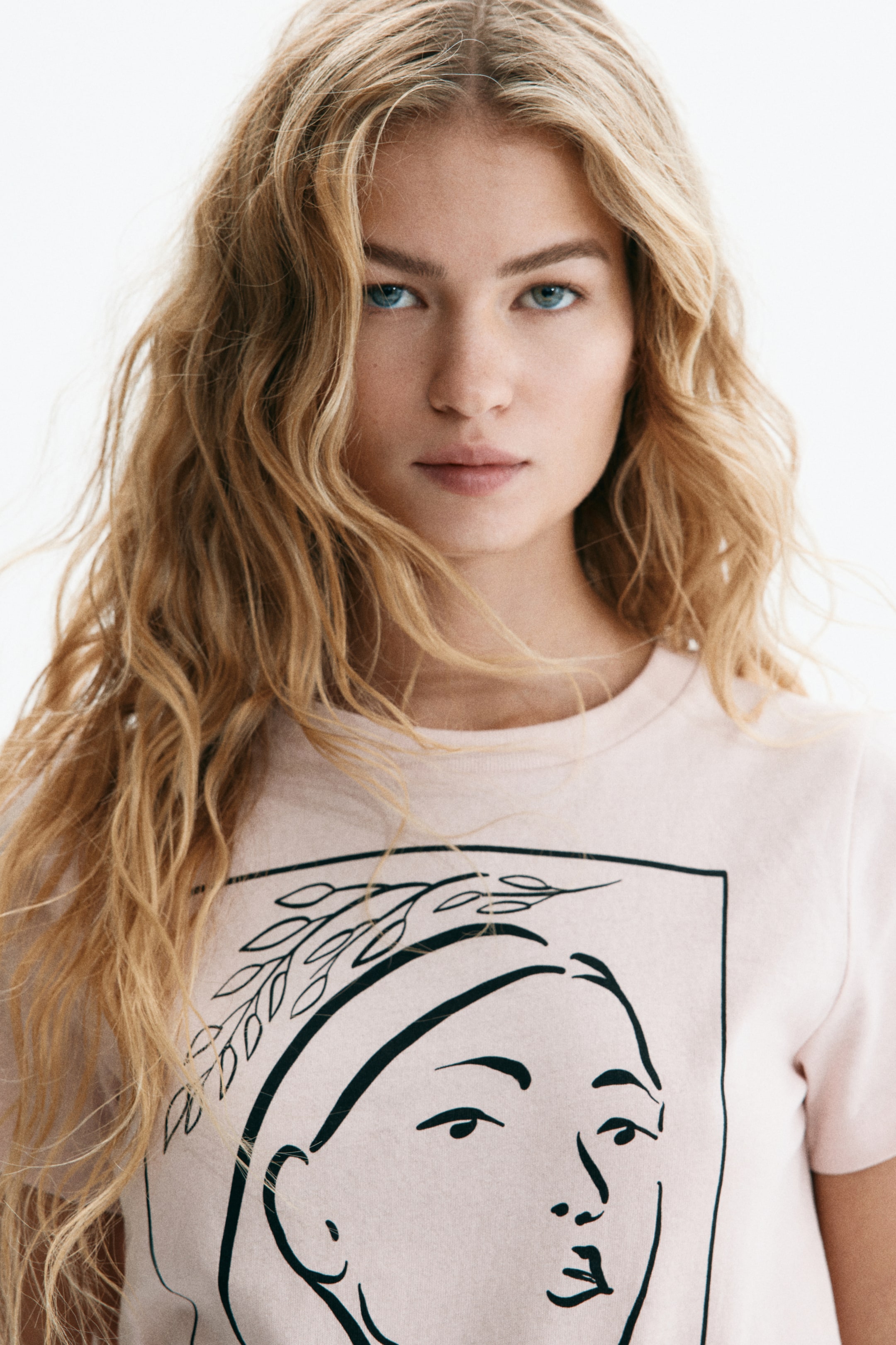 View larger image: A light-pink crew-neck T-shirt is front-facing, featuring a black line-art illustration on the front. The design depicts a stylised face in profile, facing right, enclosed within a rectangular border and adorned with a leafy wreath.
