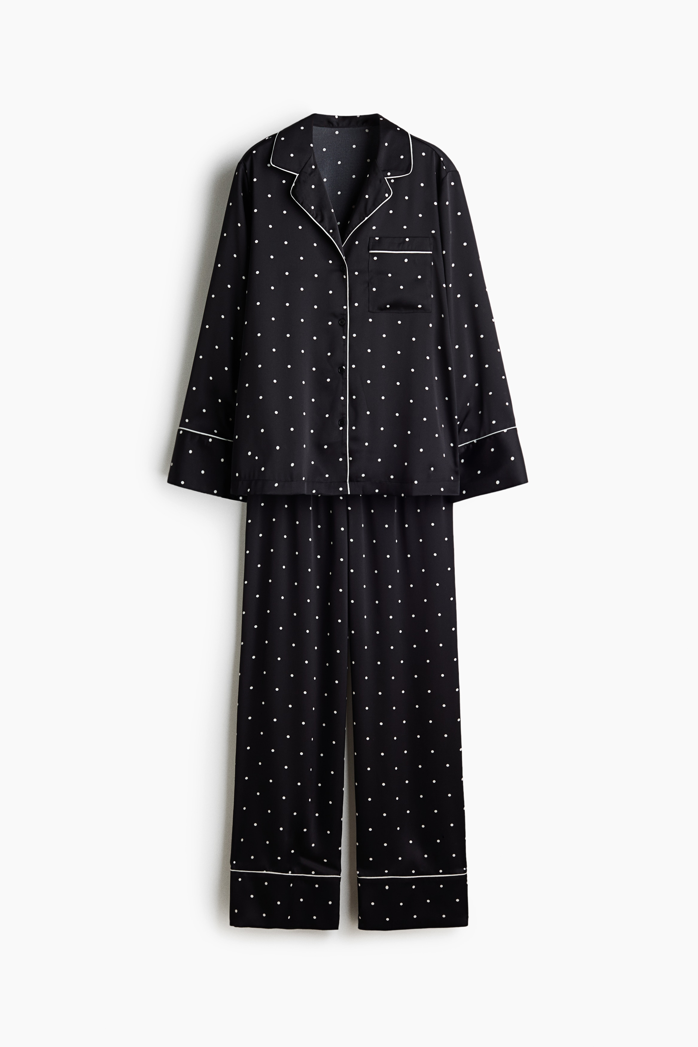 Un ensemble de pyjama en satin noir Ã  pois blancs avec passepoil blanc contrastant est portÃ© par le mannequin. La chemise Ã  manches longues prÃ©sente un col cubain, une fermeture boutonnÃ©e sur le devant et une poche poitrine, tandis que le pantalon assorti Ã  jambe large est dotÃ© dâune taille Ã  cordon de serrage visible.