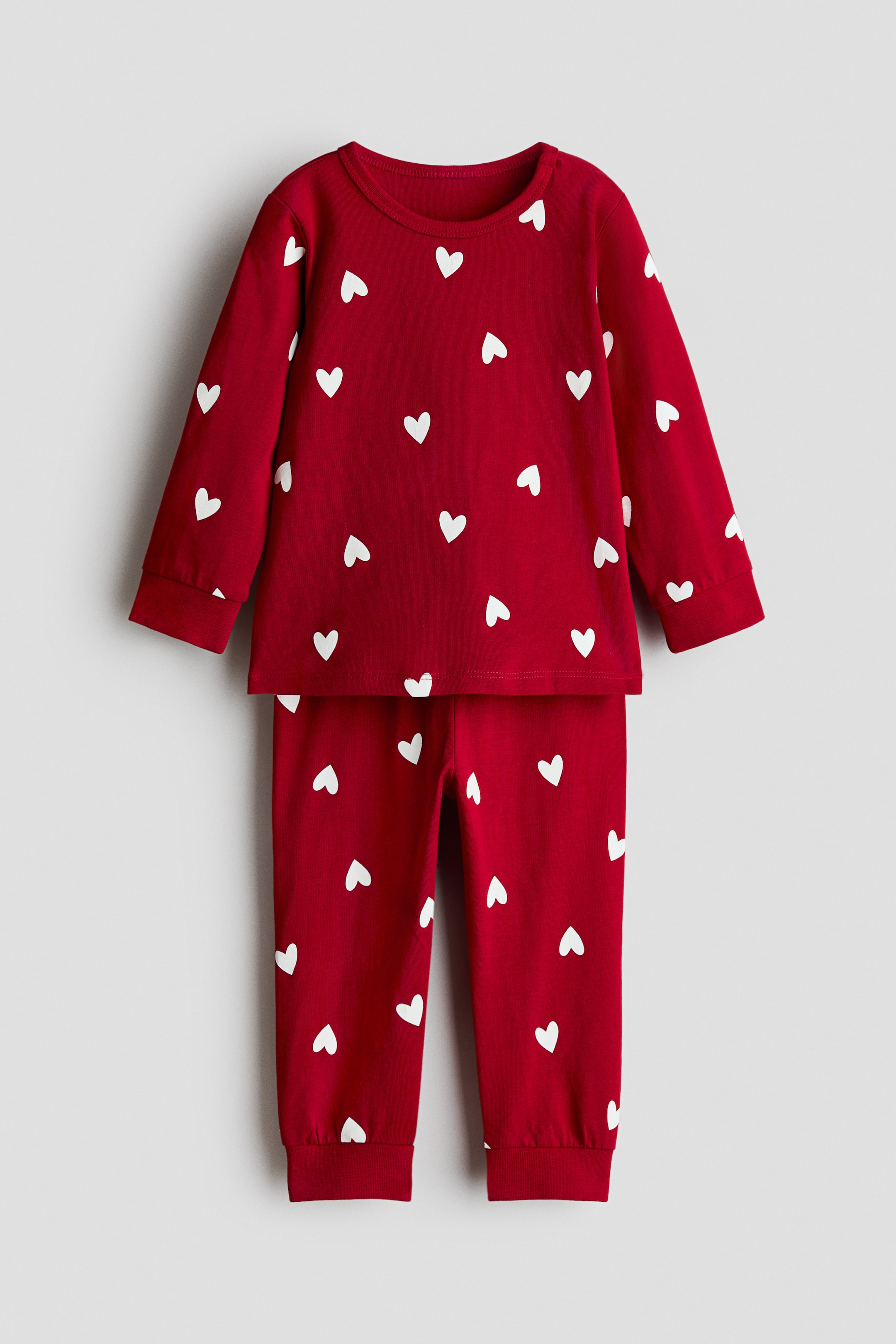 View larger image: Printed cotton pyjamas, showcased from the front, boast a rich red fabric adorned with an all-over white heart pattern. The set includes a long-sleeved top featuring a crew neck and ribbed cuffs, paired with matching long trousers that have ribbed ankle cuffs.
