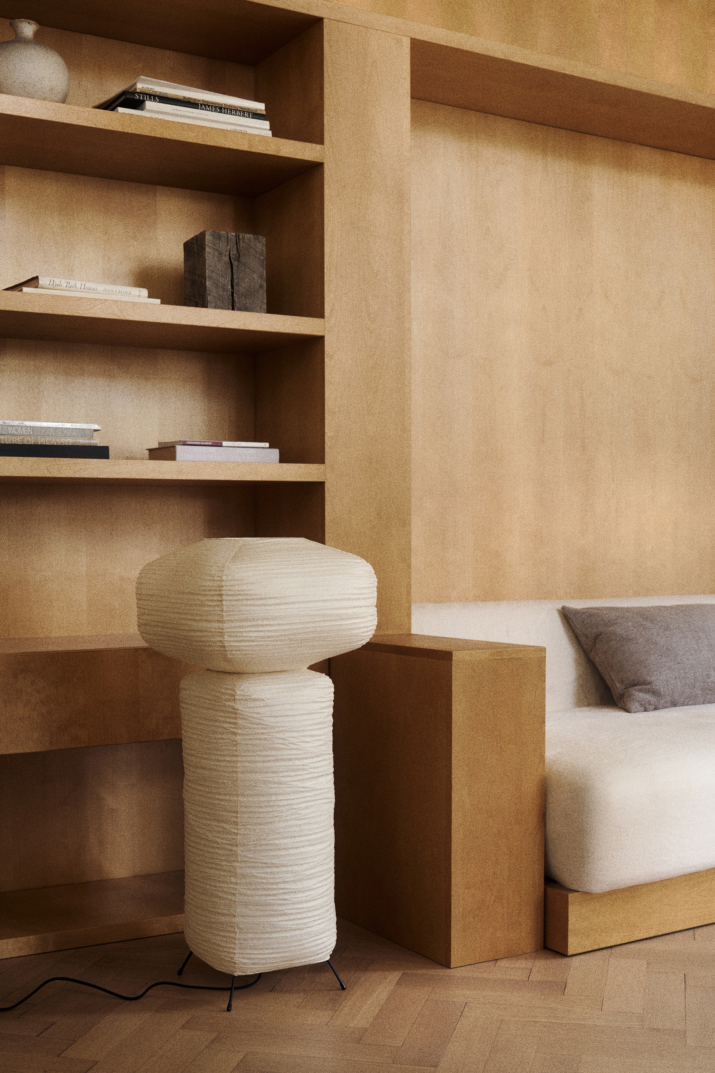 Una lámpara de pie presenta un diseño alto y apilado con dos pantallas de papel de color claro, plisadas horizontalmente. La pantalla superior tiene una forma oblonga, descansando sobre una pantalla inferior cilíndrica rectangular más alta. Está apoyada sobre una base minimalista de metal negro con tres patas extendidas, y un cable de alimentación negro se extiende desde su base.
