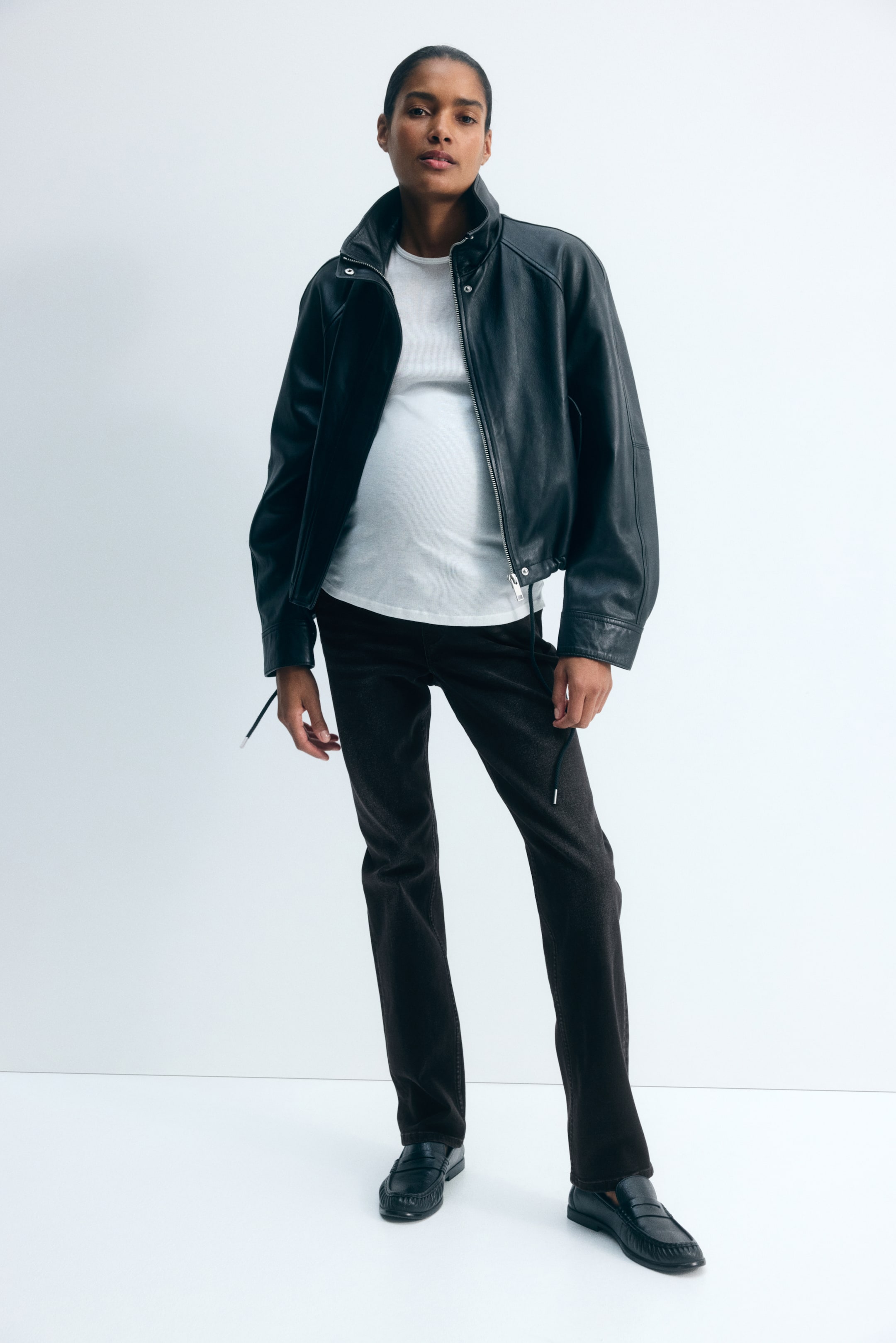 Grotere afbeelding bekijken: Donkerbruine straight-leg jeans worden gedragen door het model, met een regular fit van de dijen tot aan de zoom. De jeans worden gecombineerd met een wit T-shirt, een open zwart leren jack en zwarte loafers.
