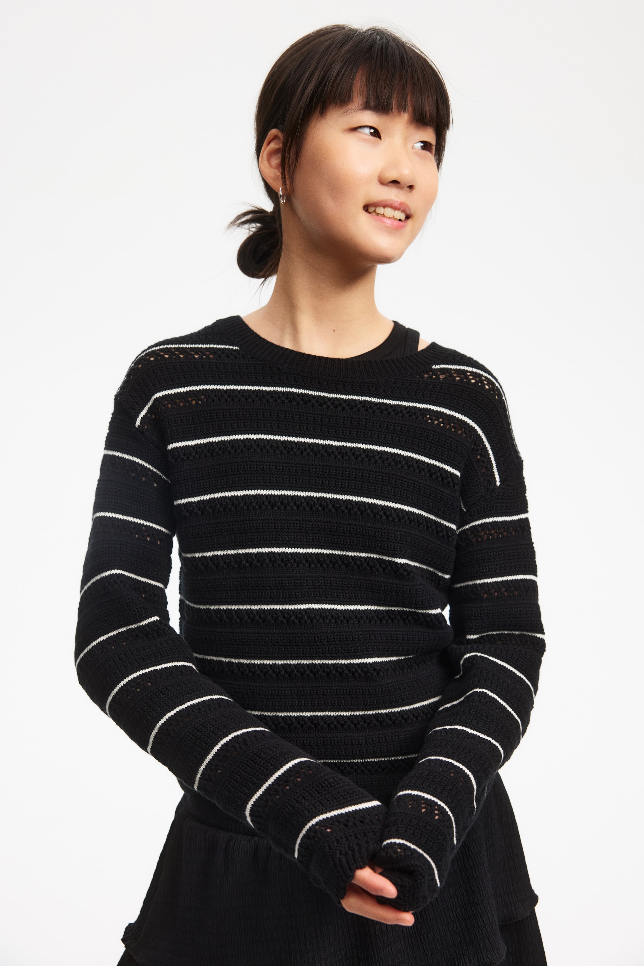 View larger image: A black open-knit sweater features horizontal white stripes across the body and long sleeves, with a dropped shoulder on the left side. A black top is visible underneath the crew neck sweater, showing through the open-knit fabric.