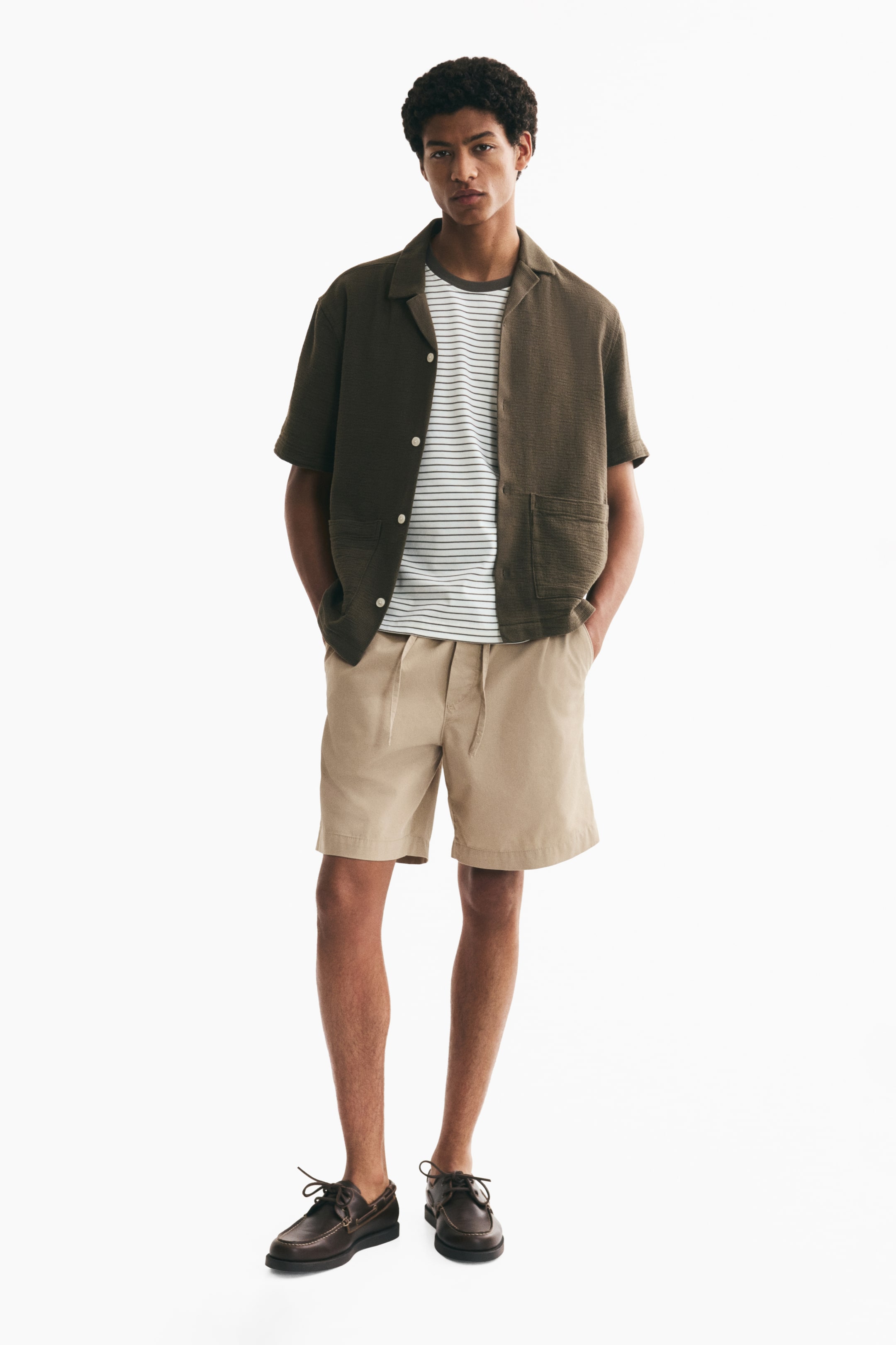 Xem ảnh lớn hơn: Mẫu đang mặc quần short vải cotton twill màu kaki. Thiết kế có cạp chun rút dây, khóa giả và túi xéo hai bên, với hai tay của mẫu đặt trong túi.