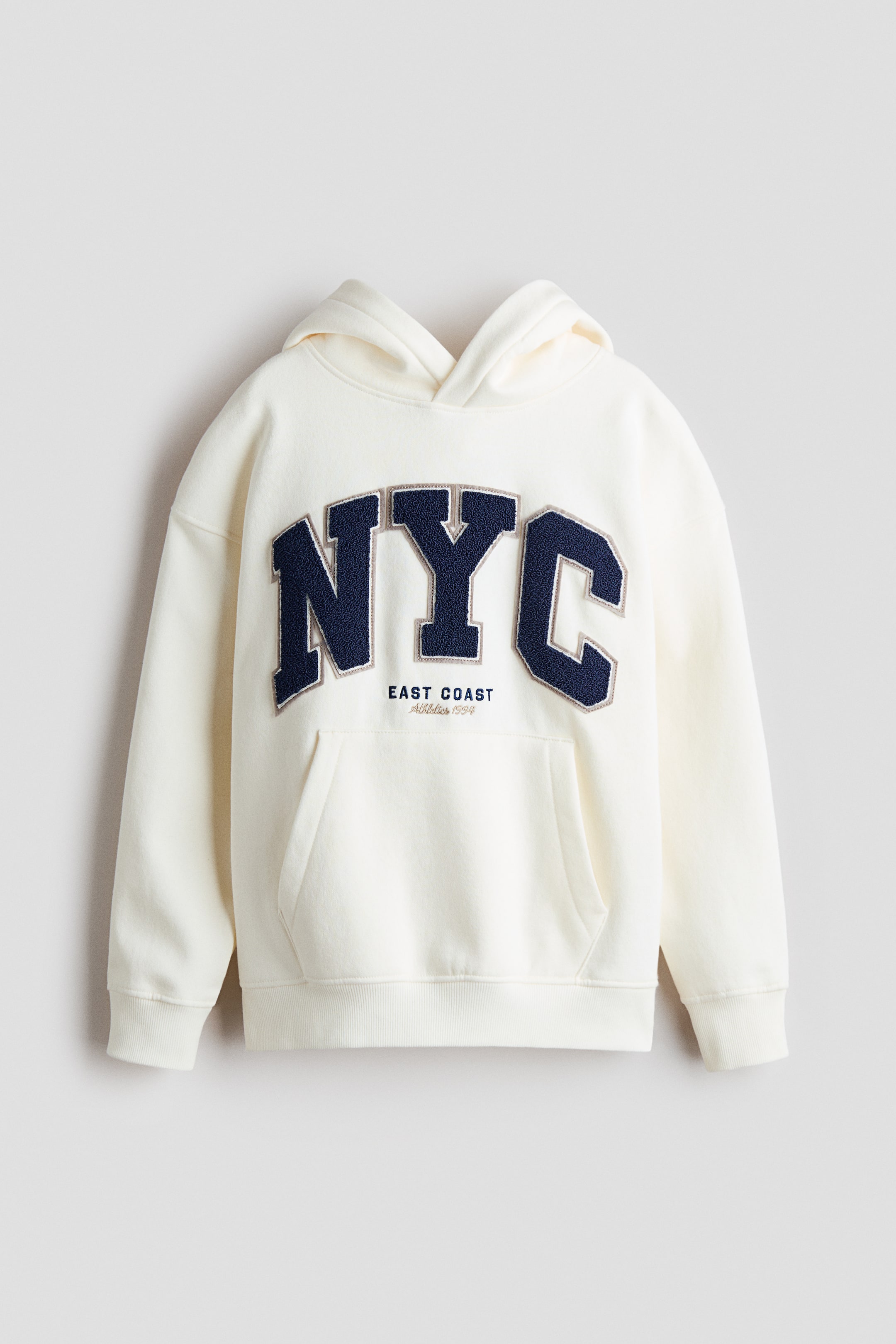 Ampliar la imagen: Una sudadera con capucha de corte holgado y color crema se presenta en el FRENTE, con una capucha de doble capa y superpuesta, hombros caídos y mangas largas. Muestra un gran bordado de letras 'NYC' en azul oscuro con un contorno claro en el pecho, encima de un texto bordado más pequeño que dice 'EAST COAST Athletics 1994', e incluye un bolsillo estilo canguro, puños acanalados y un dobladillo acanalado.