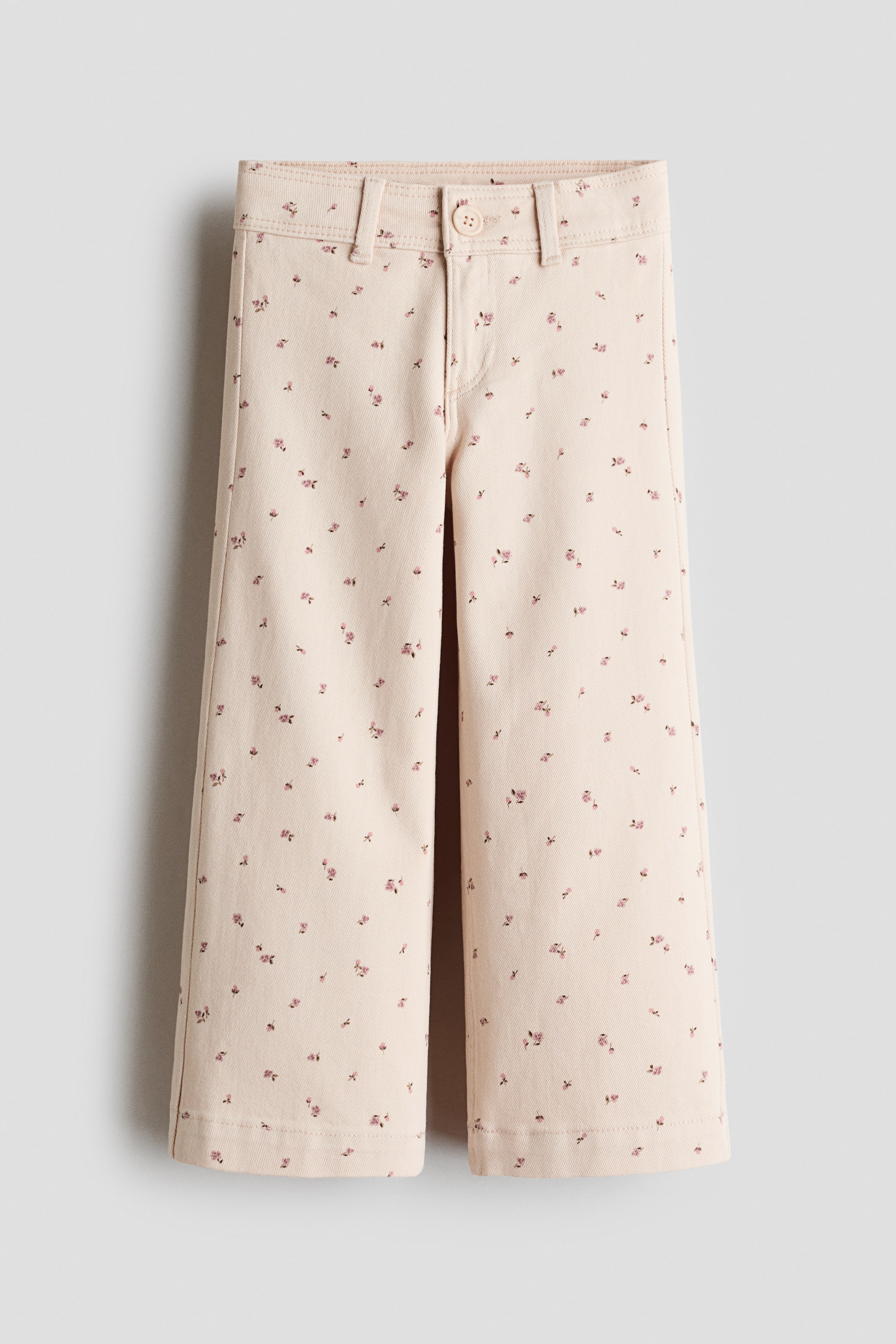 Se større billede: Lyse beige bukser vises forfra og har et spredt lyserødt blomsterprint. De er designet med en flad linning, bæltestropper og en enkel knaplukning og hænger med brede, lige ben.