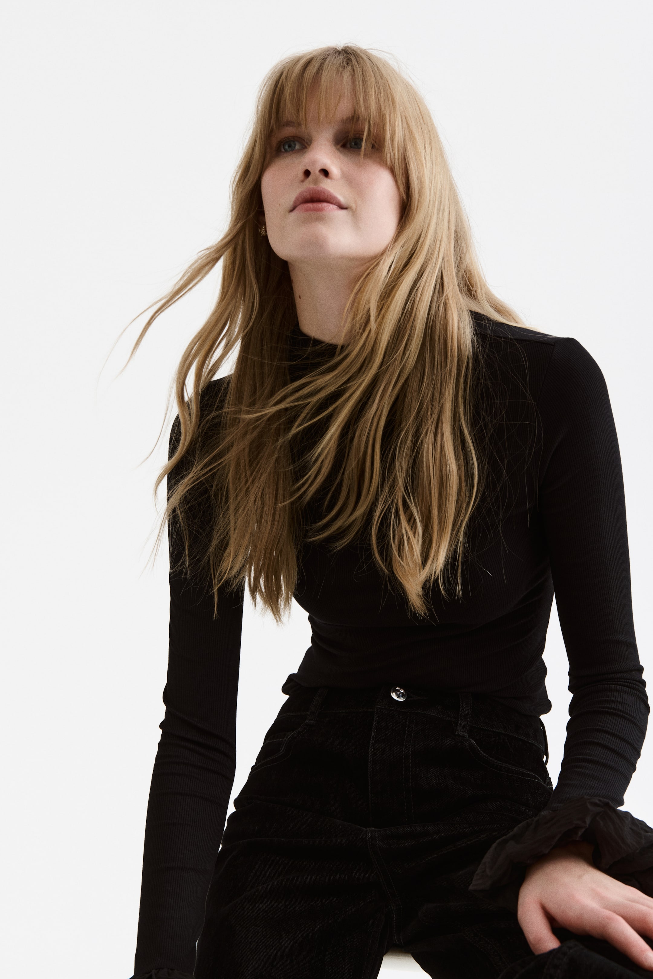 Größeres Bild anzeigen: Ein eng anliegendes, schwarzes geripptes Langarm-Top mit Stehkragen, das enge, lange Ärmel und eine figurbetonte Silhouette aufweist, wird aus einem leicht niedrigen, nach vorne gerichteten Winkel präsentiert. Es wird in schwarze Cordsamt-Hosen gesteckt getragen, die einen sichtbaren silberfarbenen Knopfverschluss und Fronttaschen zeigen.