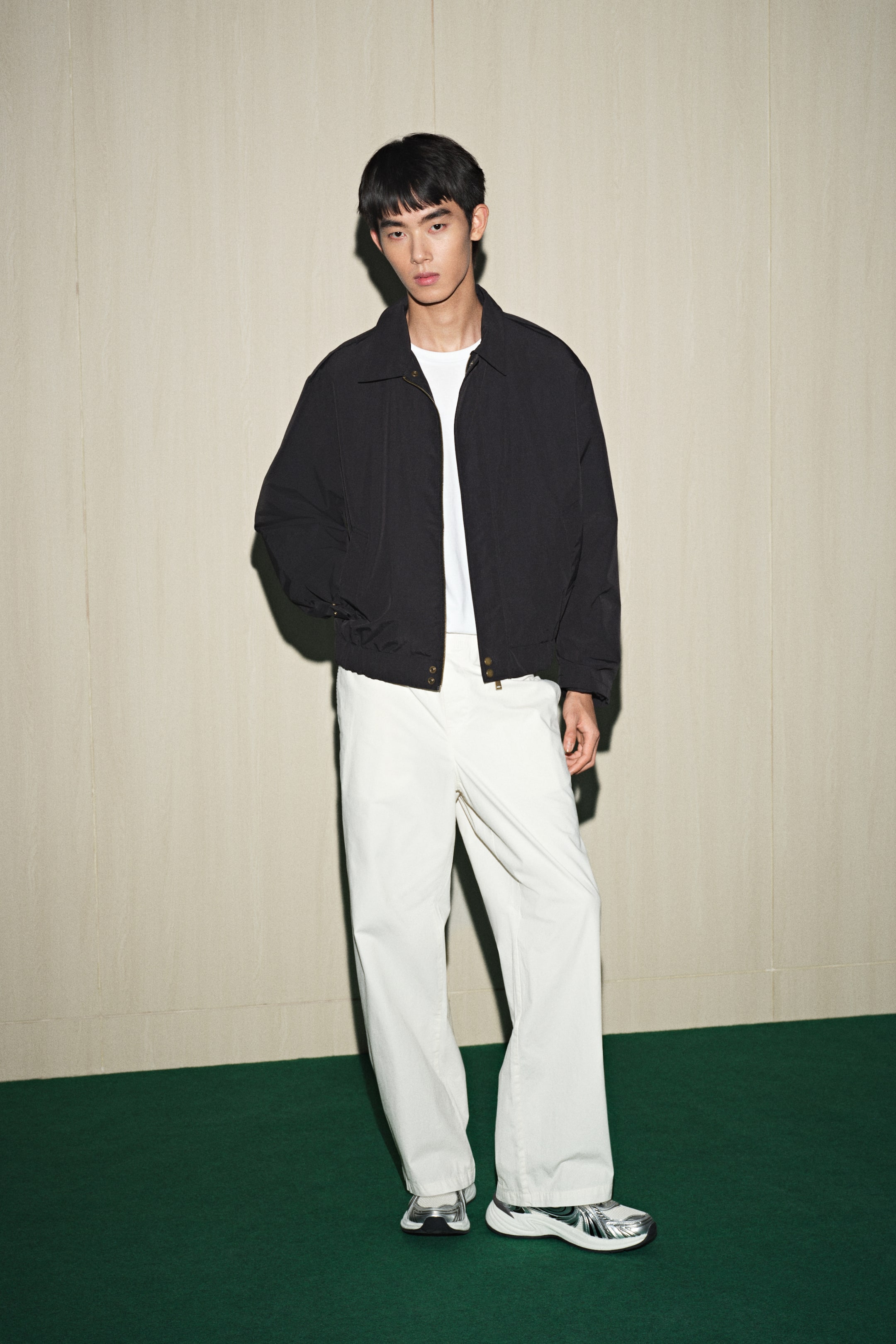 拡大画像を表示: 黒のボンバージャケットは、スプレッドカラー、フルジップクロージャー、そして伸縮性のある裾とカフスが特徴で、白のクルーネックTシャツの上に着用されています。この装いは、リラックスフィットのオフホワイトのワイドレッグパンツとシルバーのチャンキースニーカーで完成されており、すべてが前から見た状態です。