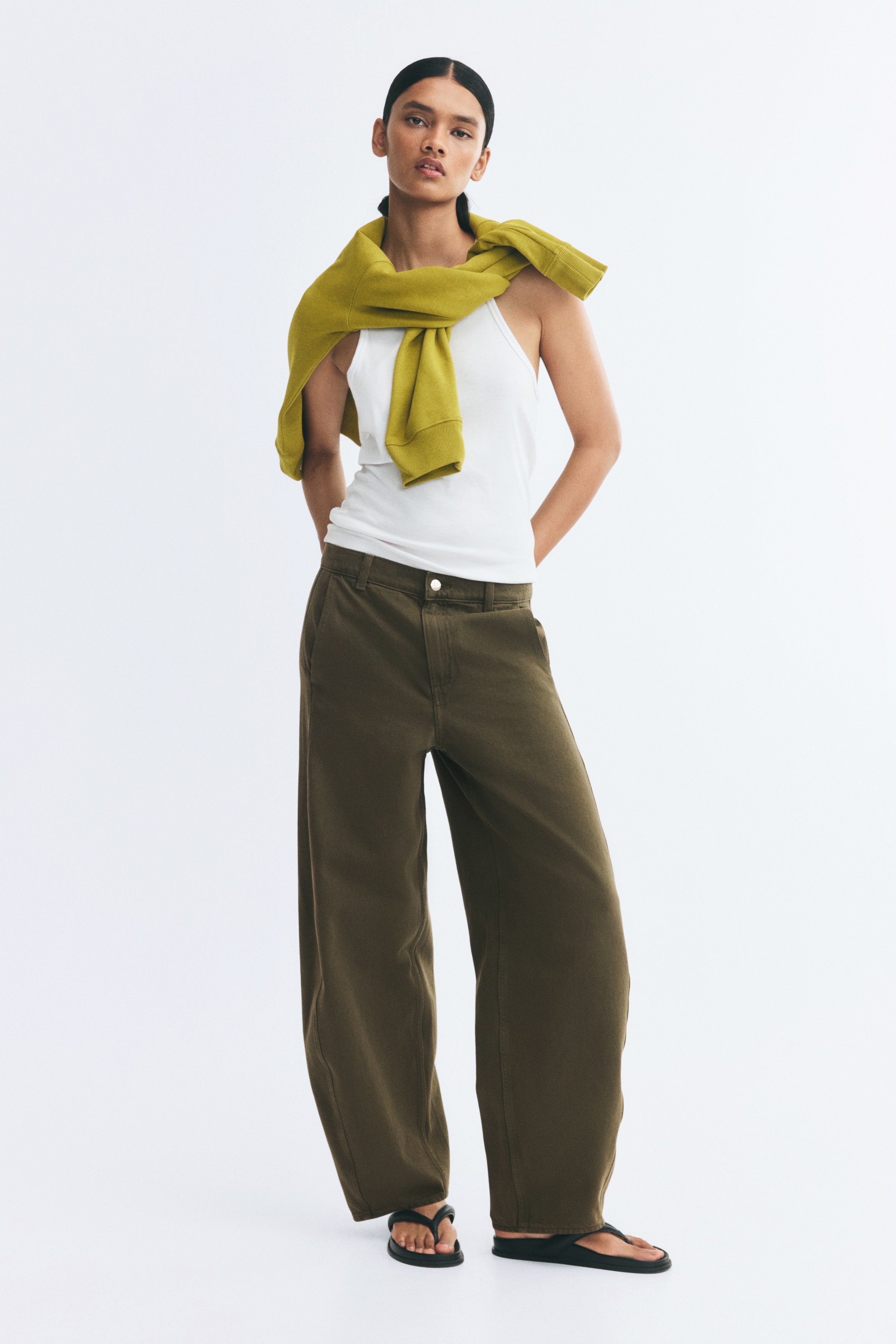 Olive brown wide-leg trousers are shown from the front, showcasing a curved barrel-leg silhouette with vertical panel seams running down each side. The trousers feature a waistband with belt loops, a silver-tone button closure, and side slant pockets.