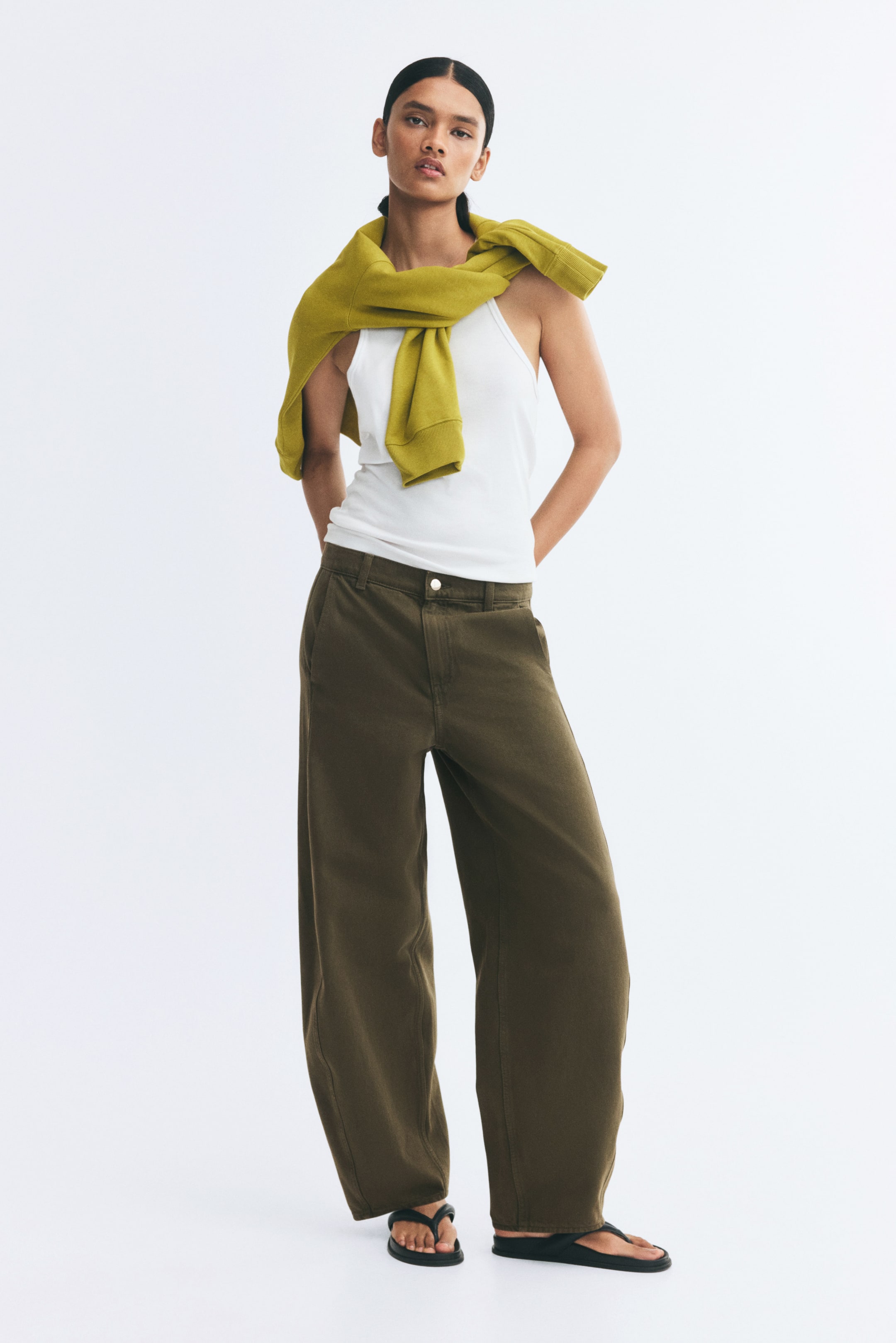 View larger image: The model is wearing olive green jeans featuring a relaxed, curved-leg silhouette and a regular waist. The tapered jeans are styled with a tucked-in white tank top and a mustard-yellow sweatshirt draped over the shoulders.