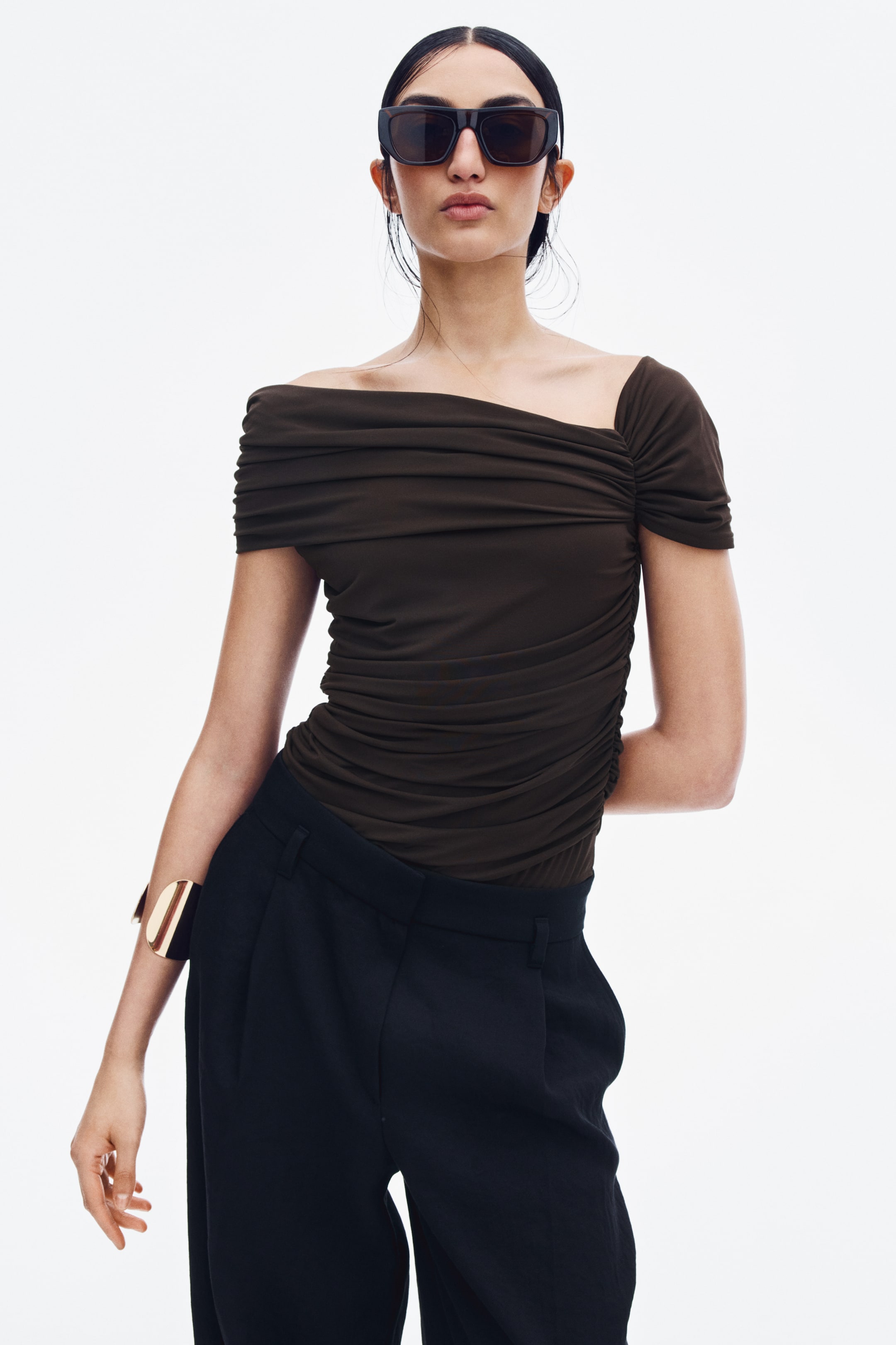 View larger image: A dark brown, asymmetrical bodysuit with horizontal gathered ruching all over is worn by the model. The off-shoulder design, featuring a draped band on one side and a short sleeve on the other, is tucked into a pair of black trousers.
