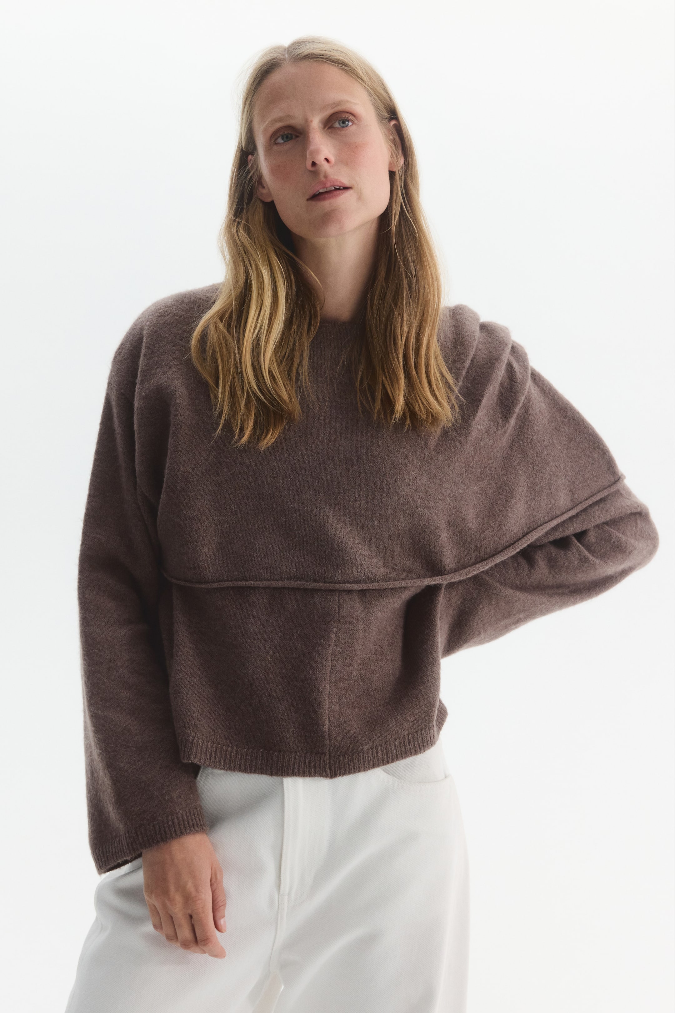 Größeres Bild anzeigen: Ein locker geschnittener brauner Strickpullover wird vom Model getragen und verfügt über einen Rundhalsausschnitt, überschnittene Schultern sowie eine asymmetrische obere Lage mit gerolltem Saum, die über eine Schulter fällt. Der Pullover wird mit einer weißen Hose kombiniert, während das Model mit einer Hand in der Hüfte posiert.