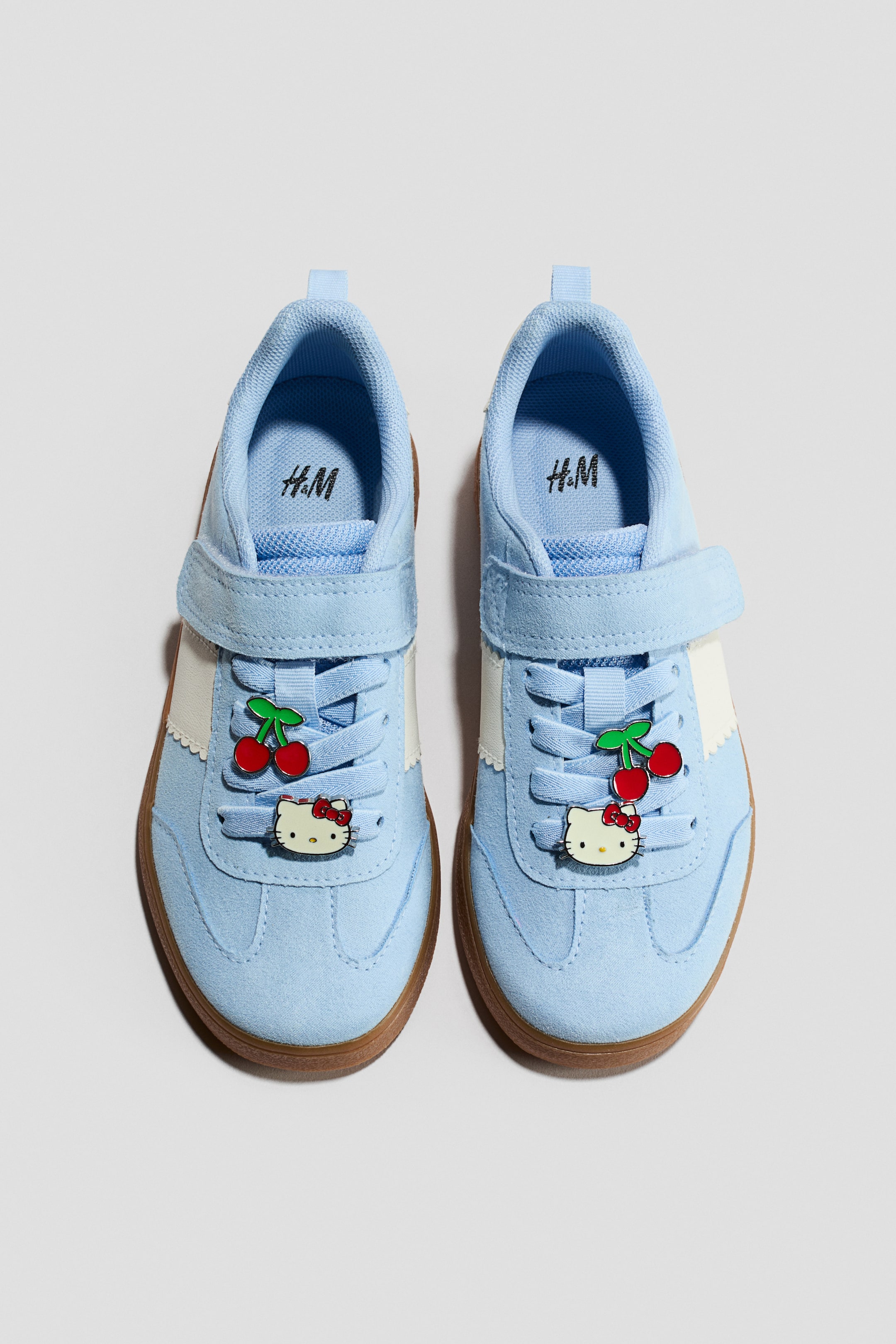 Größeres Bild anzeigen: Motiv-Detail-Sneaker. Diese hellblauen Sneaker verfügen über ein wildlederartiges Obermaterial mit weißen Seitenstreifen und einer braunen Sohle, präsentiert von der VORDERSEITE. Jeder Schuh ist mit zwei Charms verziert, einer roten Kirsche und einem weißen Hello Kitty Kopf, an den hellblauen Schnürsenkeln befestigt und durch einen Klettverschluss gesichert.