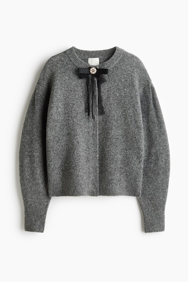A grey marled knit cardigan is shown facing front, featuring a ribbed crew neck, dropped shoulders, and long ribbed cuffs and hem. The cardigan is decorated with a black ribbon bow at the neckline, accented with a pearl and rhinestone flower embellishment. - 1