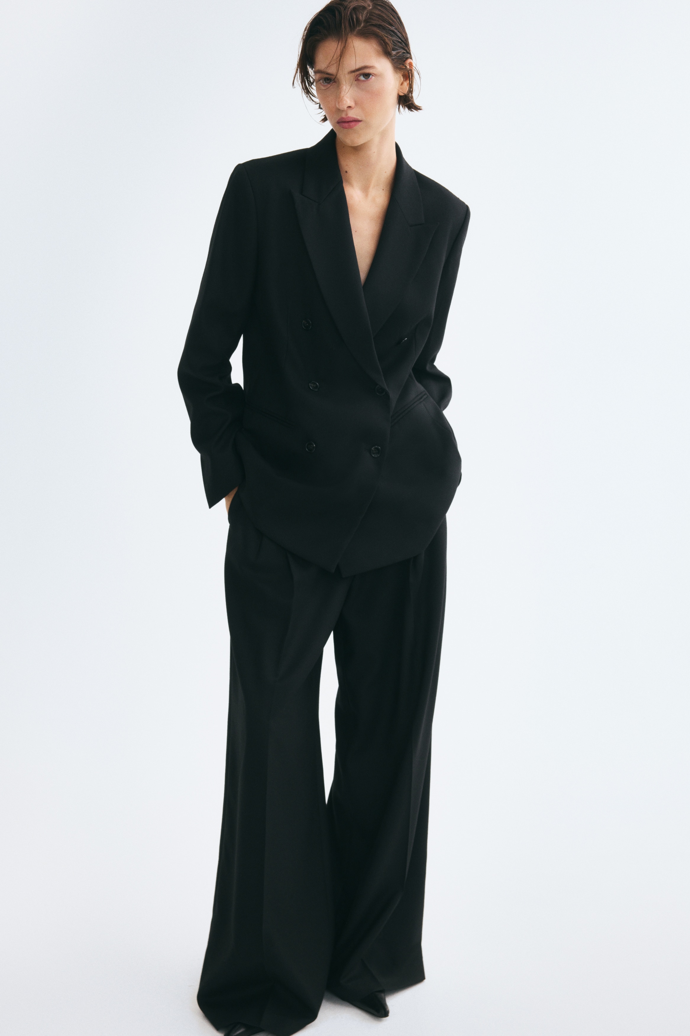 A black, double-breasted blazer is shown from the front, featuring wide pointed lapels, a six-button front fastening, and welt front pockets. The structured design showcases padded shoulders and tailored waist darts.