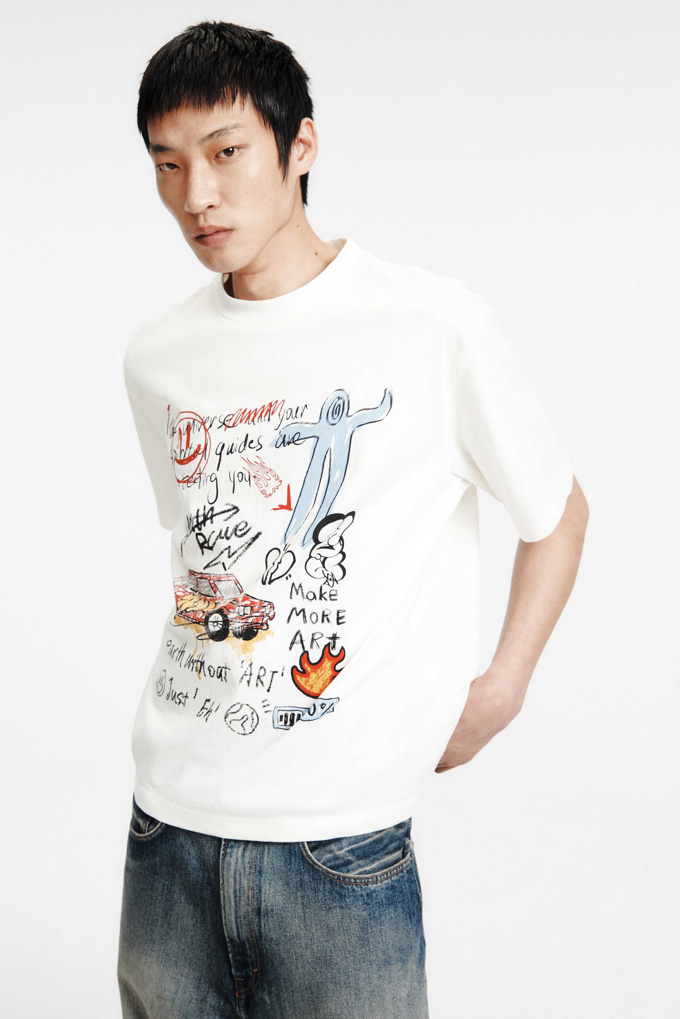 拡大画像を表示: オフホワイトのTシャツは、リブ付きのクルーネックとショートスリーブが特徴で、フロントには手描き風のスタイル要素を含むマルチカラーのグラフィックプリントが施されています。要素にはテキスト、車、人物、炎が含まれています。青いジーンズの上にゆったりと垂れ下がり、裾が見えています。