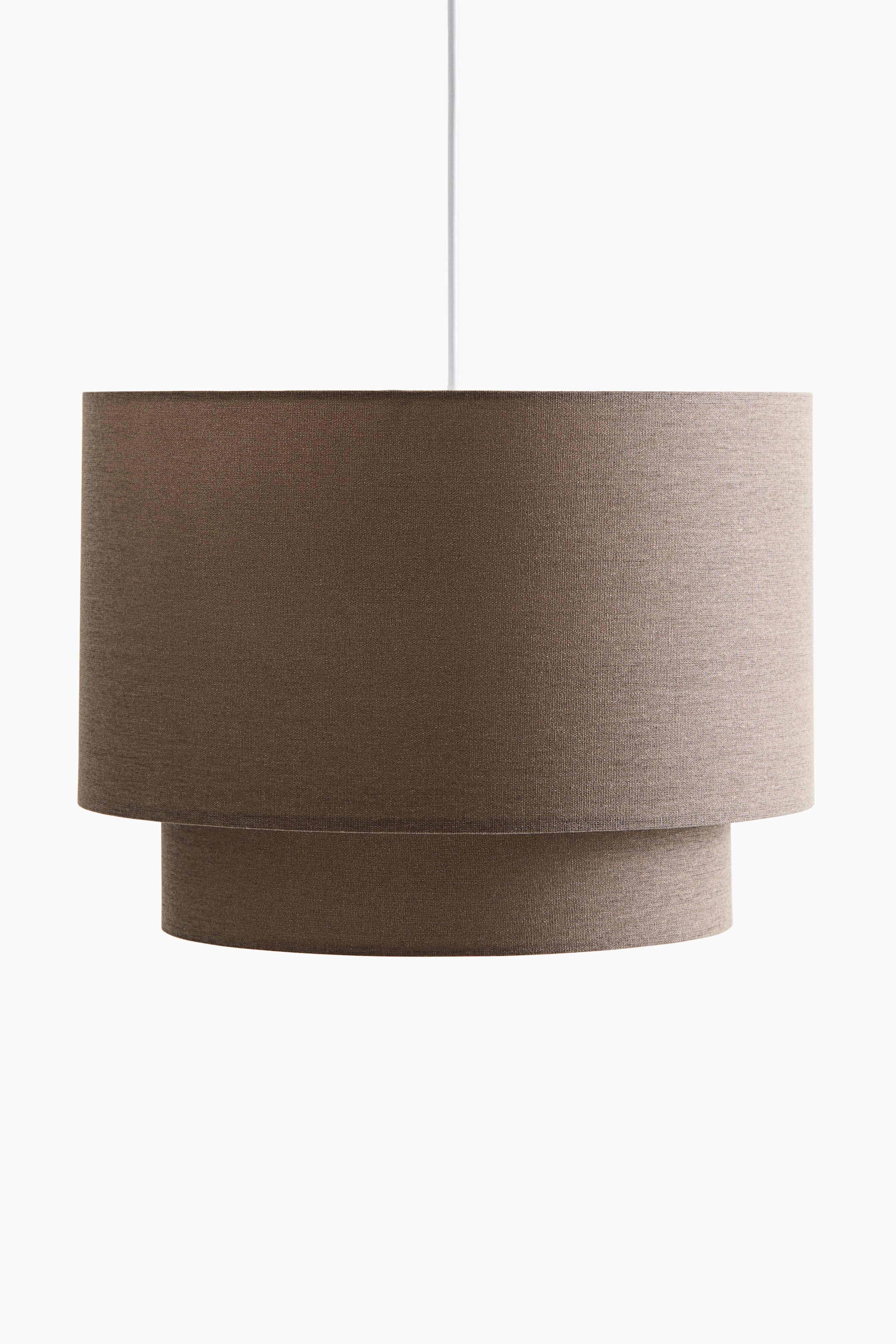 View larger image: A pendant light, facing front, features two layered cylindrical shades crafted from neutral brown fabric. The upper shade appears wider than the lower, with a white cord suspending the light.