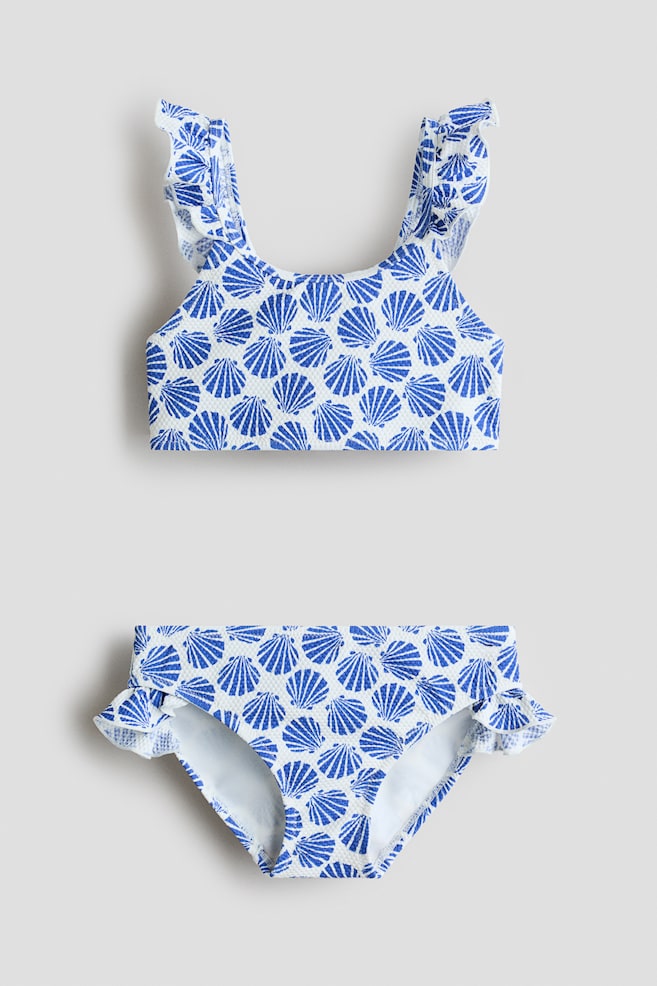 O biquíni de duas peças está deitado plano, virado para a frente, exibindo um tecido texturizado branco com um padrão de conchas azuis. O top apresenta alças com folhos nos ombros, e a parte de baixo exibe detalhes franzidos nas laterais. - 1