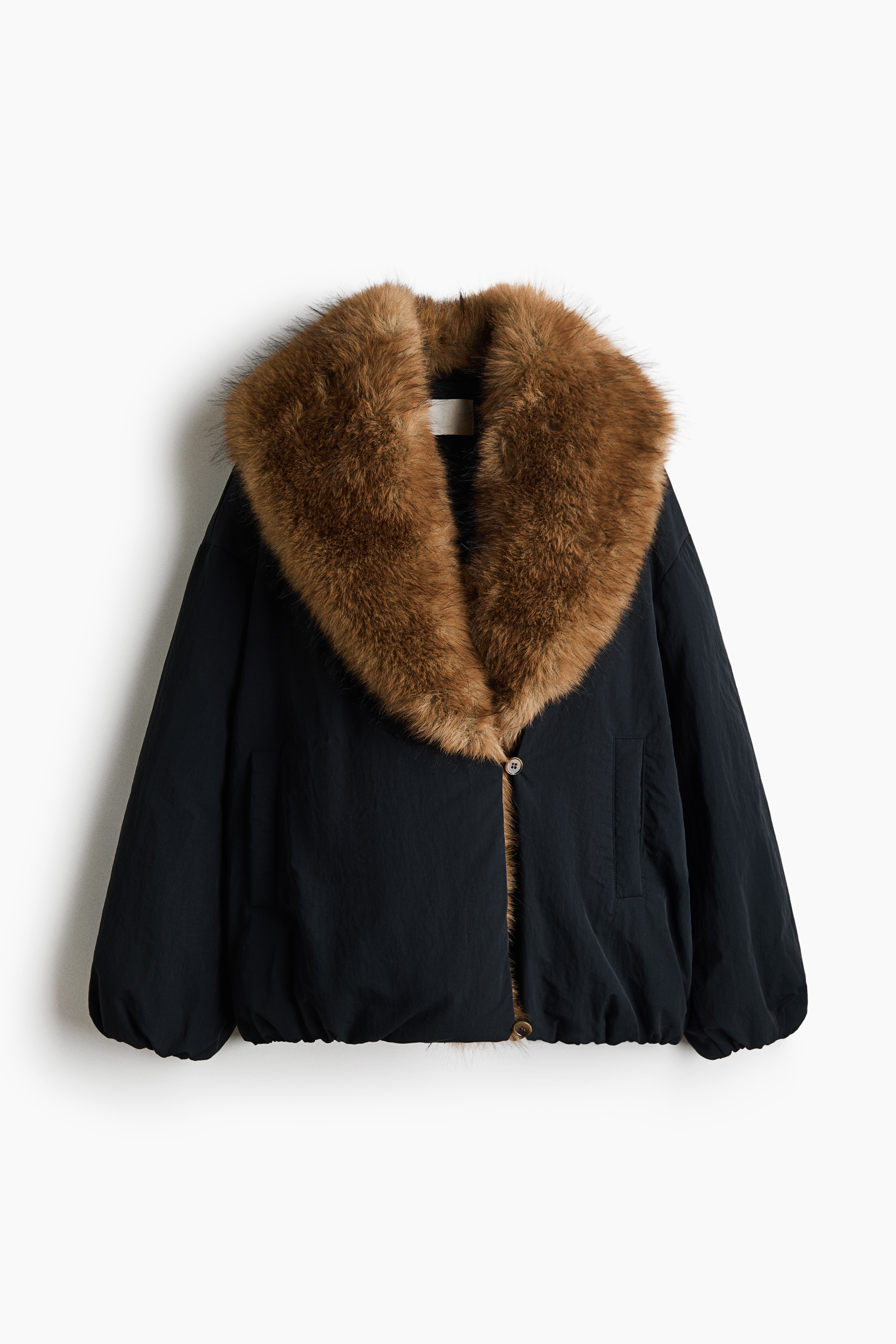 An oversized, padded black nylon jacket featuring a wide brown faux-fur shawl collar is being worn by the model. The model is holding a black leather handbag with silver buckles against the front of the jacket, which is styled with dark leather trousers.