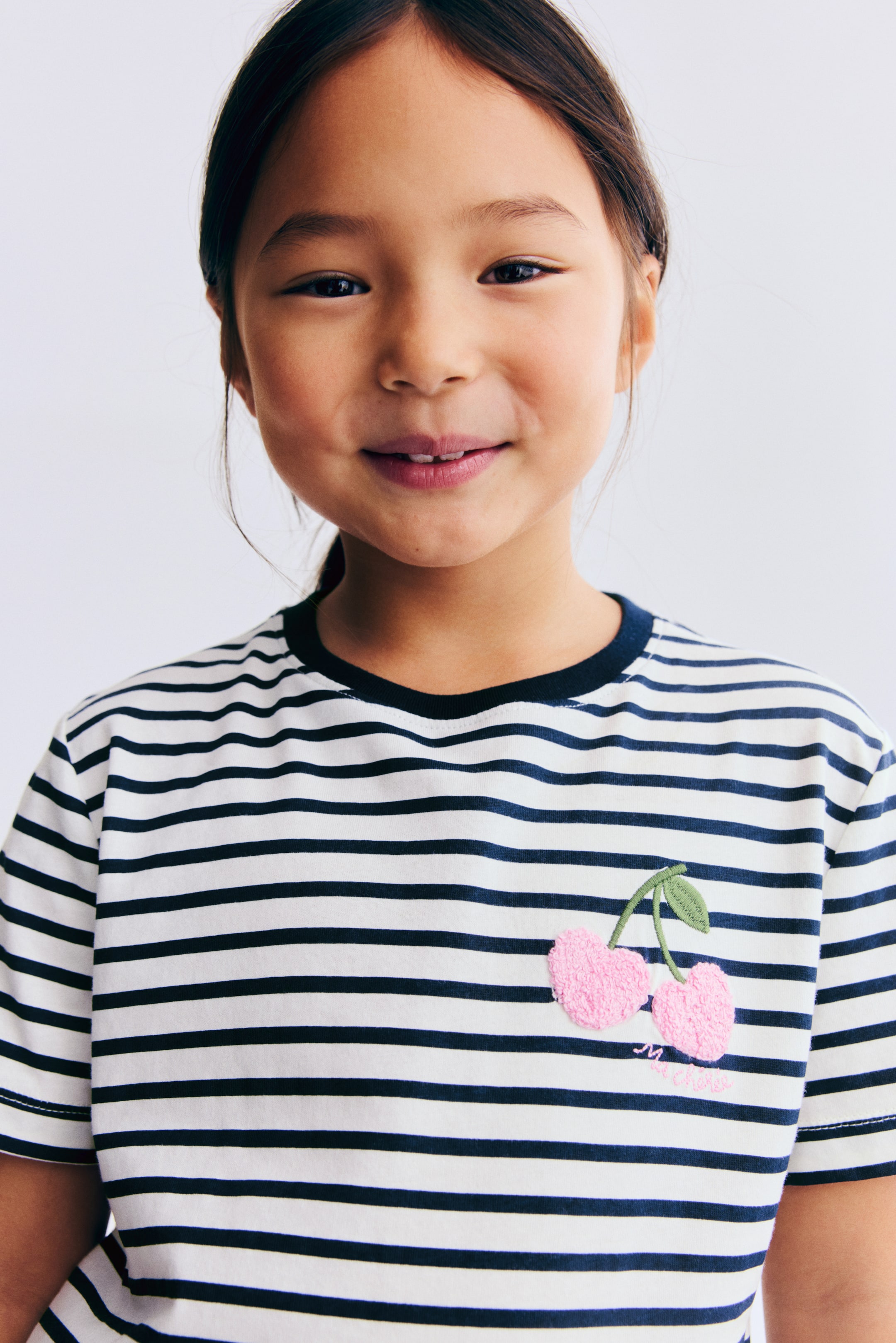 הצגת תמונה גדולה יותר: החולצה מעוצבת בפסים אופקיים לבנים וכחול נייבי. היא מציגה צווארון עגול ריב בכחול נייבי, ועיטור רקום של שתי דובדבנים ורודים במרקם, עם גבעול ועלה ירוק, מקשט את החלק העליון השמאלי של החזית באזור החזה.