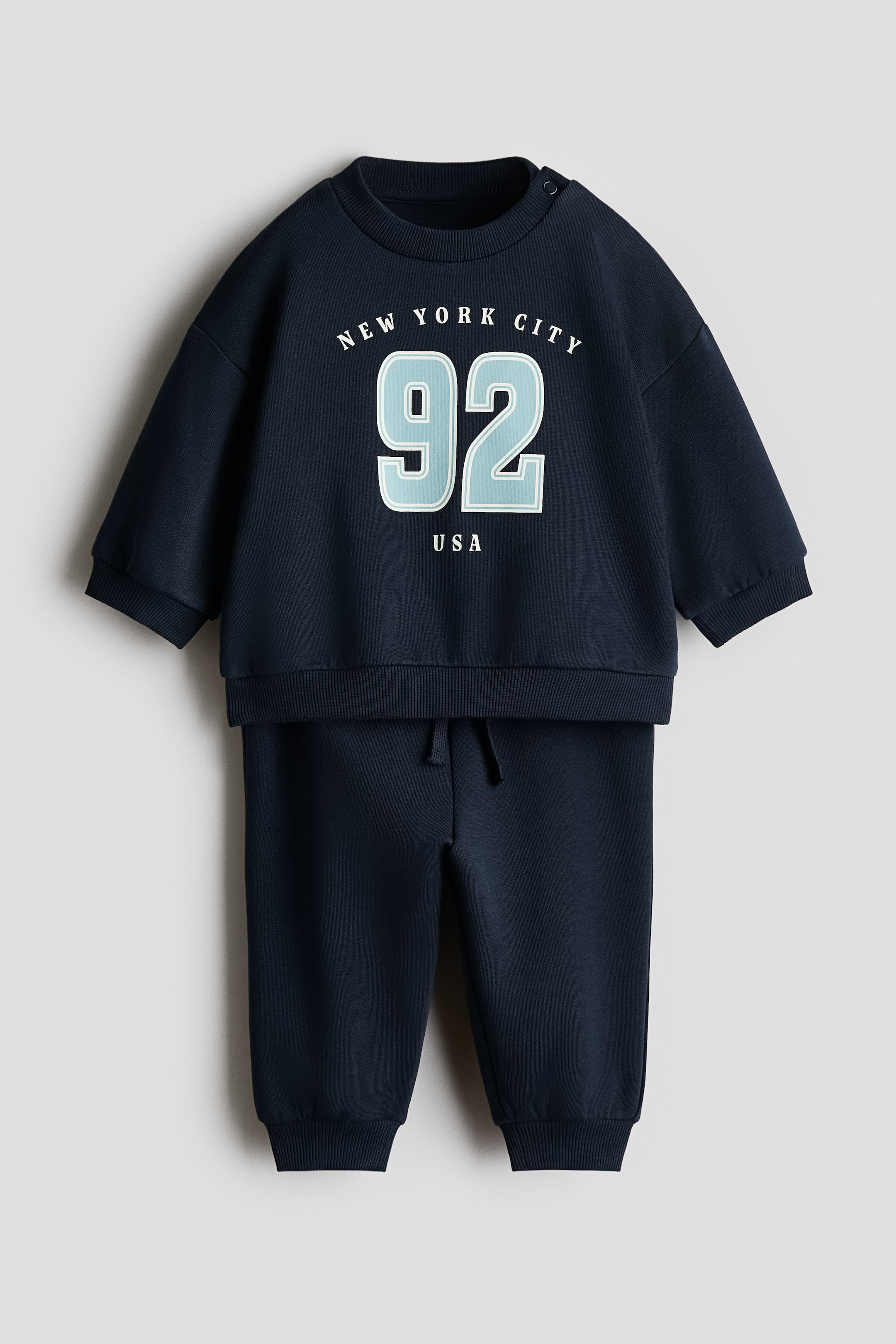 View larger image: A navy blue baby tracksuit set comprising a sweatshirt and matching joggers, presented front-facing. The sweatshirt features a "NEW YORK CITY 92 USA" graphic print on the front, a crew neck, ribbed cuffs, a ribbed hem, and two snap buttons on the left shoulder. The joggers showcase an elastic waistband with a drawstring and ribbed ankle cuffs.