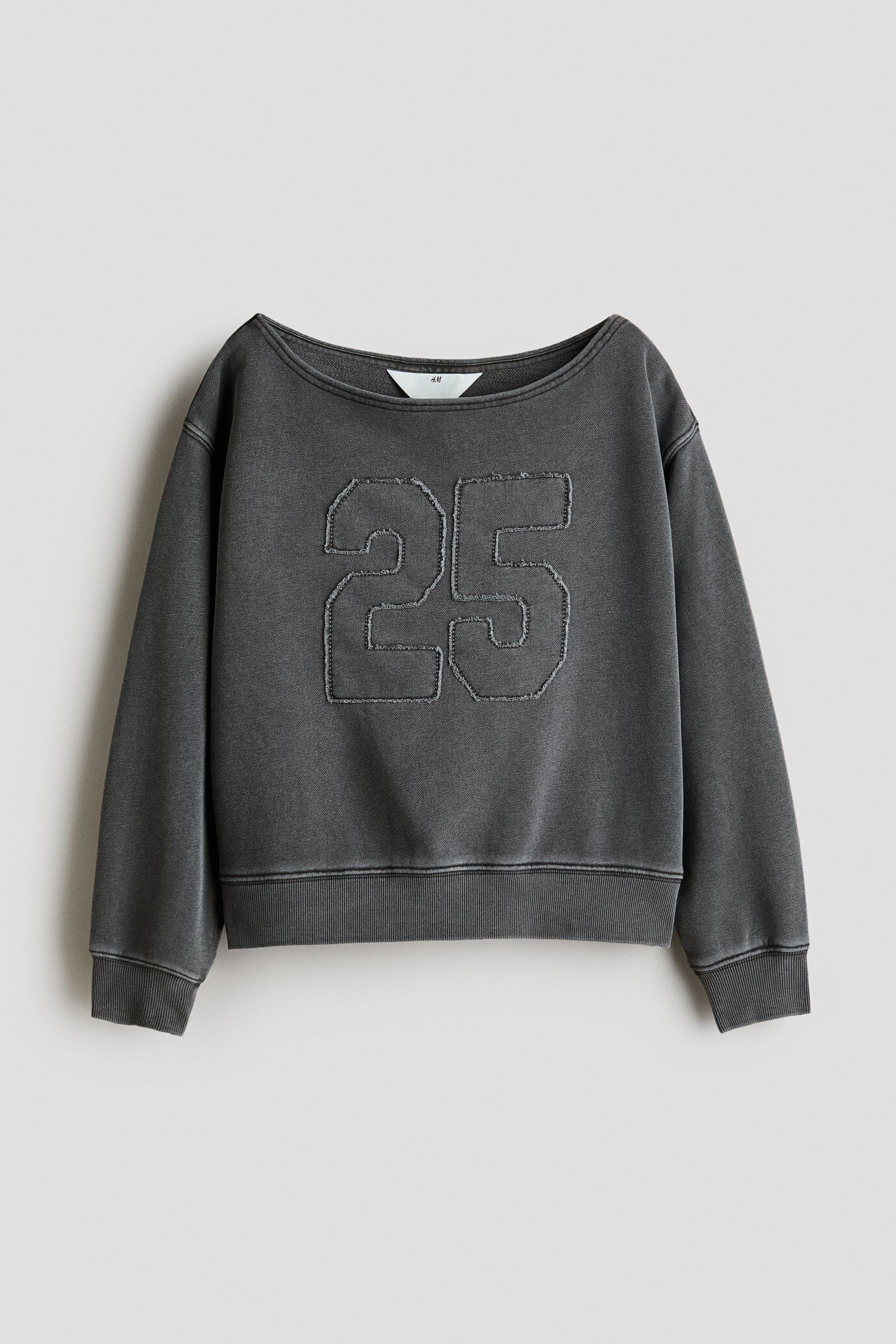 Ampliar la imagen: Una sudadera gris oscuro se presenta de FRENTE, con un amplio escote barco y mangas largas con puños acanalados. Un gran aplique '25' con bordes desgastados está colocado centralmente en el frente, y también presenta un dobladillo acanalado.