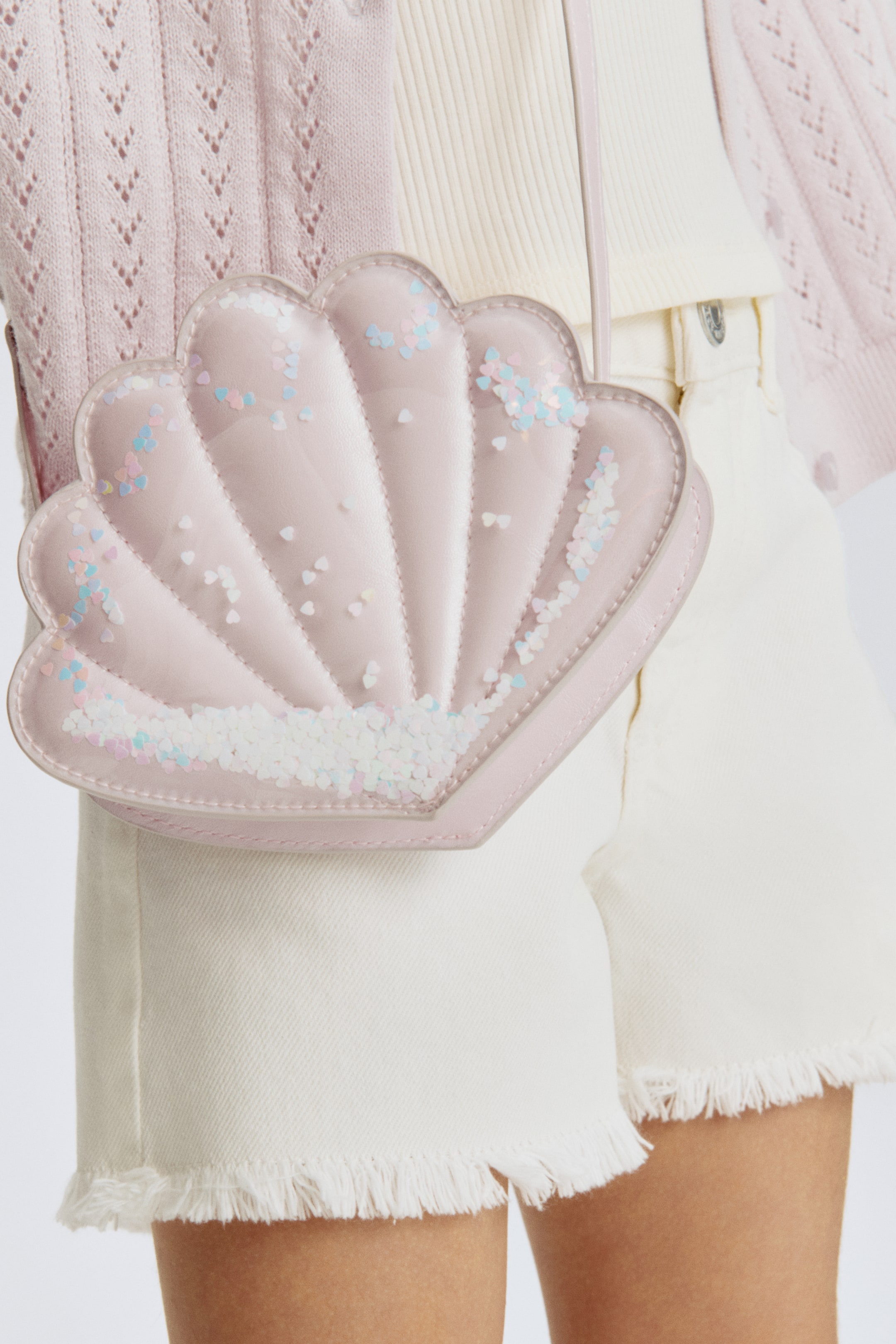 Visa större bild: En framåtvänd, snäckformad crossbody-väska i ljusrosa, skimrande, vadderat tyg med räfflade sektioner. Ett klart överdrag täcker framsidan och innehåller spridda hjärtformade konfetti i rosa, blått och vitt, med en smal ljusrosa axelrem som sträcker sig från den övre kanten.