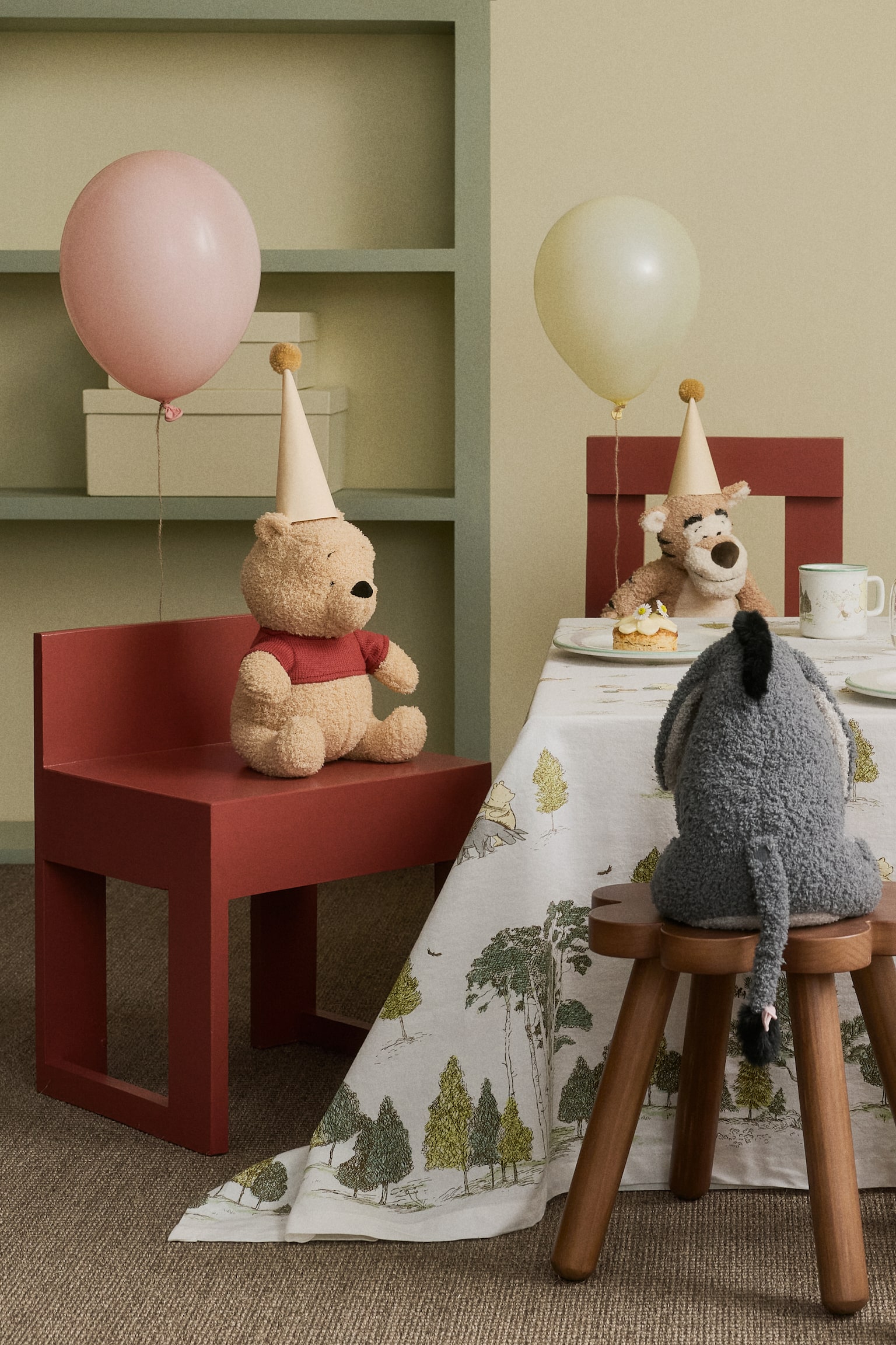 Ein Stofftier, gestaltet als Winnie Puuh, sitzt leicht nach rechts gewandt. Es besteht aus hellbraunem Teddystoff mit gestickten Gesichtszügen, trägt ein rotes kurzärmliges Hemd und einen hellbeigen konischen Partyhut mit einem gelben Pompon. - 1